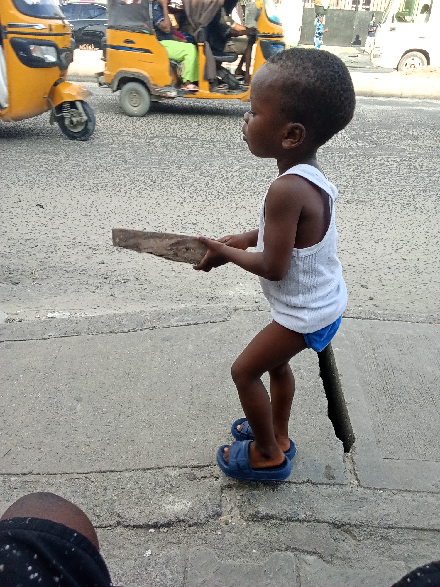 A young Black child, likely a boy, stands on a cracked concrete sidewalk in Ado, Nigeria. The child is wearing a white tank top and blue underpants, with blue slide sandals on their feet. In their hands, they hold a flat piece of wood. Behind the child, a busy street is visible with several motorized rickshaws, commonly known as "keke napeps," which are a popular mode of transport in Nigeria. Some people are visible riding in or near the rickshaws. The background suggests an urban environment with buildings and the general hustle and bustle of city life. The child appears to be standing on a raised section of the sidewalk, with a large crack in the concrete.