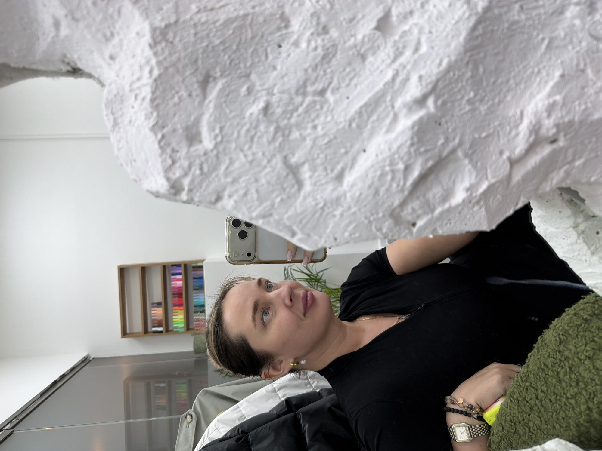 A woman is taking a selfie in a room. She is holding a phone in her right hand, and her face is visible in the frame. The background includes a white wall, a shelf with colorful items, and a mirror. The location cannot be determined from the image.