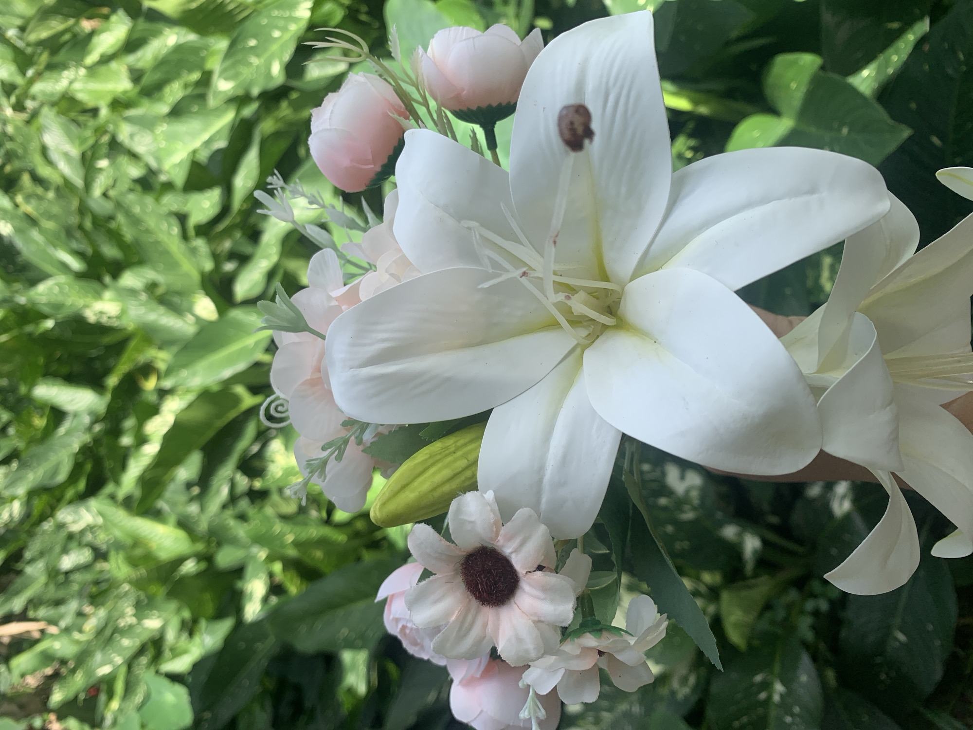A close-up, vertical shot shows a bouquet of artificial flowers held by a hand against a blurred backdrop of lush green foliage. The bouquet prominently features a large, white lily with yellow accents in its center and a small, brown spot on one of its petals. Surrounding the lily are smaller, pale pink roses, a light pink daisy with a dark center, and a pale green bud. The foliage in the background appears to be large-leafed plants, possibly in a garden or park. The overall impression is one of a decorative arrangement presented in a natural setting. There is no specific information within the image to indicate a location in Kuala Pembuang, Indonesia.