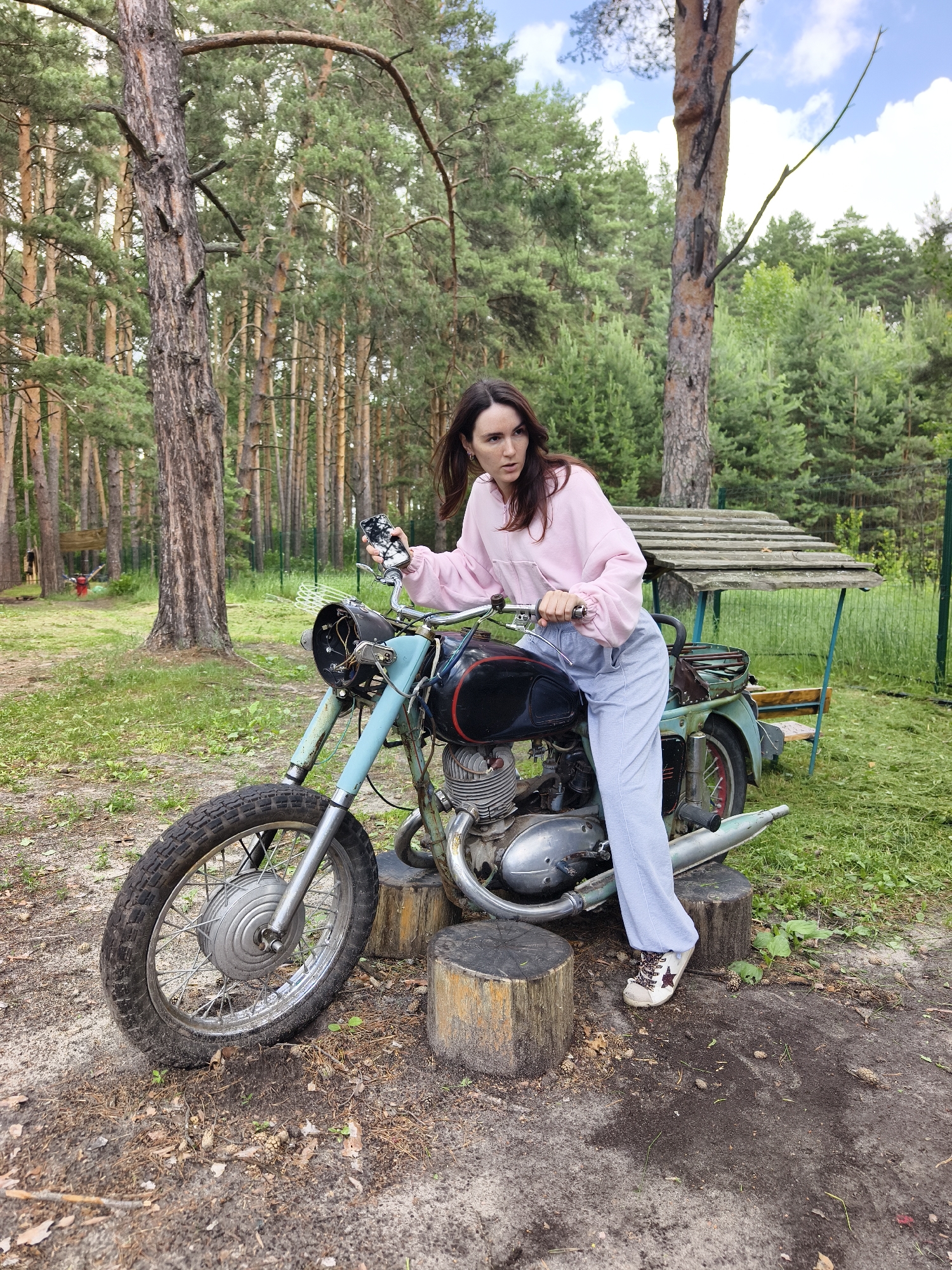 A young woman with long brown hair is sitting on an old, light blue and black motorcycle in a forest clearing. She is wearing a pink hoodie and light blue pants with a striped pattern. Her right hand is on the handlebar, and her left hand is holding a smartphone. The motorcycle is resting on two wooden stumps. In the background, there are tall pine trees, green grass, and a rustic wooden shelter. The ground is covered with pine needles and dirt.