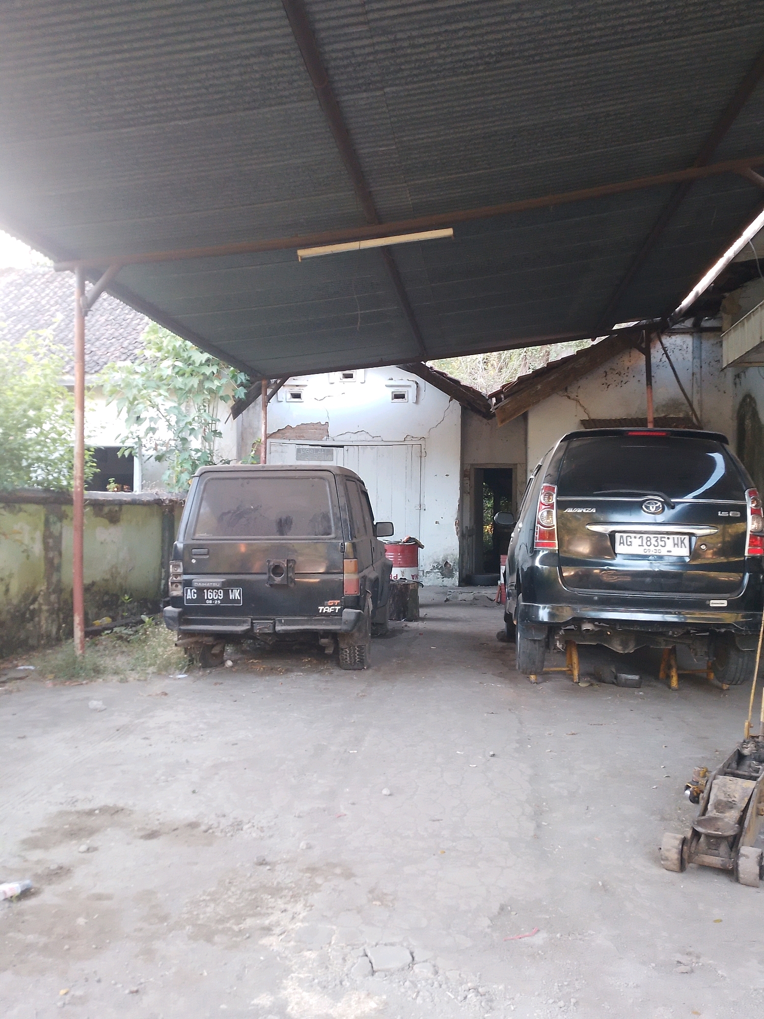 The image shows a dusty, outdoor car repair area in Patianrowo, Indonesia. Two black vehicles are parked side-by-side under a corrugated metal roof supported by metal poles. The vehicle on the left is a Daihatsu Taft, with license plate "AG 1669 WK". The vehicle on the right is a Toyota Avanza, with license plate "AG 1835 WK". The Avanza is raised on yellow support stands. A yellow hydraulic jack is visible in the foreground to the right. The ground is unpaved and appears to be dirt and gravel, with some patches of mud. A partially visible building with white walls and a wooden door is in the background. Overgrown vegetation is present to the left of the vehicles. The lighting suggests it is daytime, possibly late afternoon.