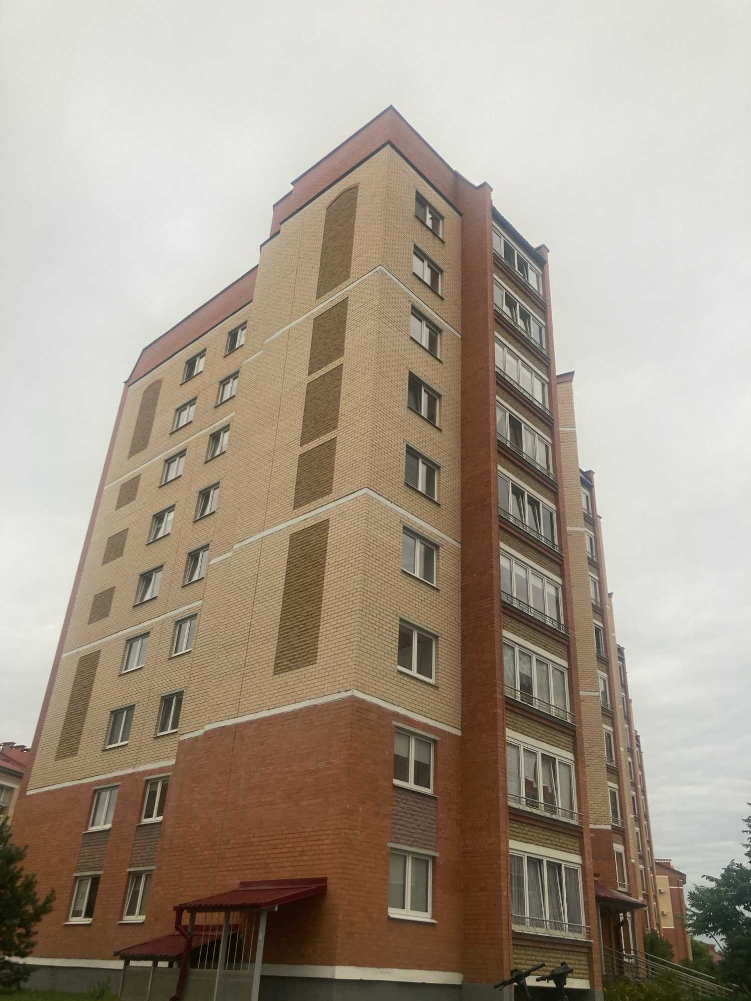 A low-angle, full shot captures a multi-story brick apartment building against a cloudy sky. The building features a light tan brick facade with darker tan rectangular accents arranged vertically and horizontally. The lower portion of the building is clad in dark red brick. The building has numerous windows with white frames, some of which are open. A red awning shades an entrance with a metal railing. The image appears to be taken in the city of Brest, Belarus.