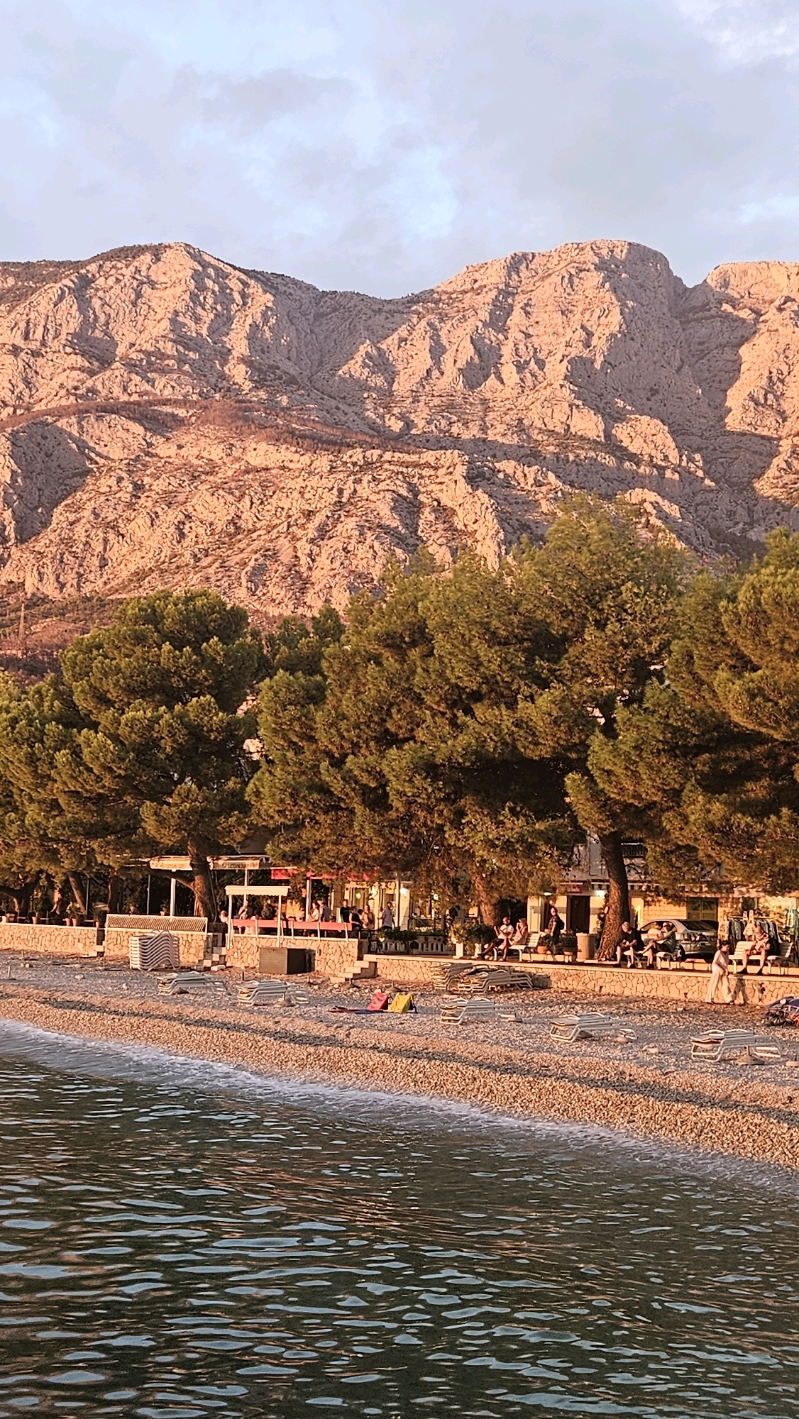 The image depicts a seaside scene at sunset in Općina Tučepi, Croatia. A pebble beach is in the foreground, with the gentle waves of the Adriatic Sea lapping at the shore. Beyond the beach, a line of pine trees provides shade for a row of cafes and restaurants. People are seen seated at tables, enjoying the evening atmosphere. In the background, a majestic mountain range rises, its rocky slopes bathed in the warm, golden light of the setting sun. The sky above is a soft blue, with scattered clouds. The overall impression is one of a tranquil and picturesque coastal setting.