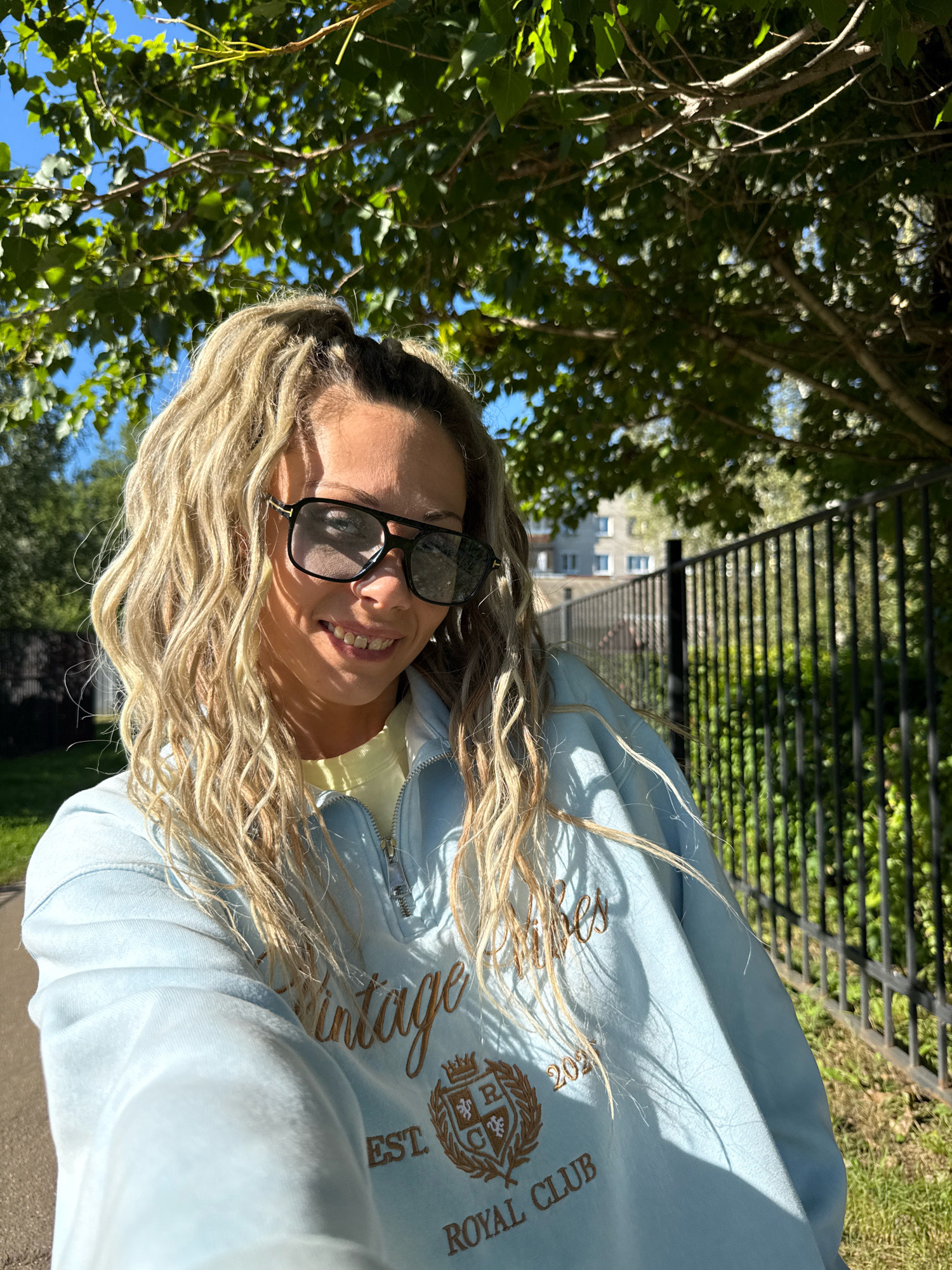 A young woman with blonde, wavy hair pulled into a high ponytail and wearing large black sunglasses is taking a selfie outdoors in what appears to be a park or residential area. She is smiling and wearing a light blue quarter-zip sweatshirt with "Vintage Royal Club Est. 2021" embroidered on the front. The background shows a black metal fence, green bushes and grass, a tree with green leaves and sunlight filtering through, and a building with windows in the distance. The sun is shining, casting shadows on the woman and the ground. The location is specified as Zhukovo, Russia.