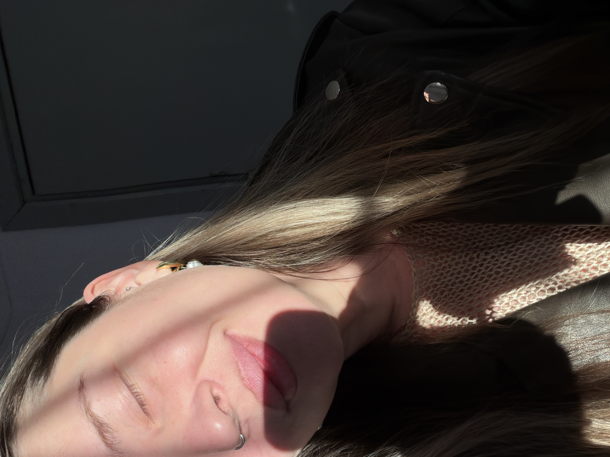 A close-up, tilted, and sunlit view features a fair-skinned person with closed eyes. The individual has blonde-tinted brown hair, a nose ring, and an earring. The person appears to be wearing a beige netted garment. A black garment with shiny silver buttons is partially visible. Part of a dark grey wall is visible in the background. The image was taken indoors, based on the lighting and visible interior elements. The location is not discernible based on visual evidence.