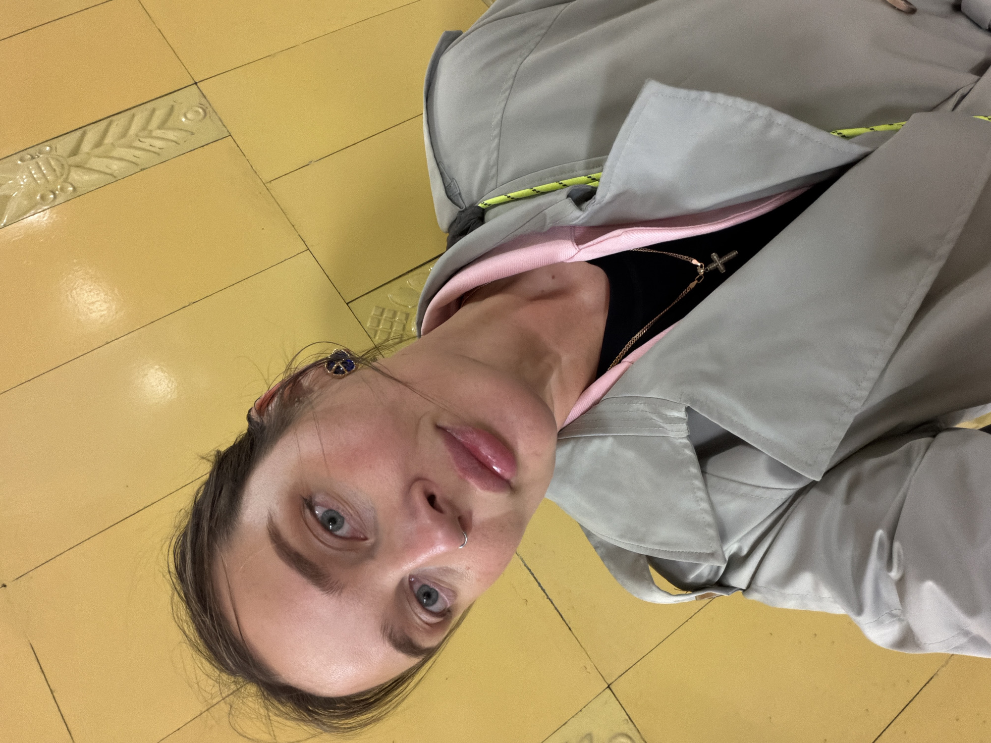 A person lies on a yellow tiled floor. The tiles have an ornamental pattern along the edges. The person is wearing a gray coat over a pink hoodie and a black top, with a gold chain and cross visible. They have a nose ring and an earring. The location is likely a subway station in Moscow, Russia, indicated by the distinct yellow tile pattern of certain stations.