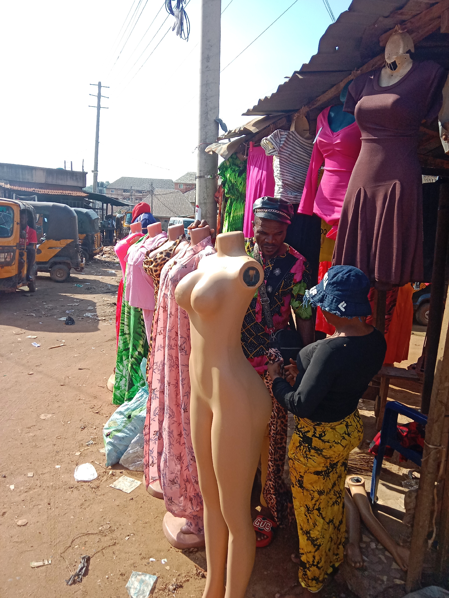 The image depicts an outdoor clothing stall in what appears to be a market area in Nkpor, Nigeria. Two people are present. A man wearing a black leather cap and a colorful floral shirt is seen interacting with a woman wearing a black long-sleeved shirt, a yellow patterned skirt, and a blue denim bucket hat. They are examining a piece of clothing. The stall displays a variety of dresses on mannequins, including pink, green, leopard print, and a solid brown dress. A headless mannequin is standing prominently at the front. Behind the stall, several yellow auto-rickshaws (tuk-tuks) are parked along the street, and buildings can be seen in the background. The ground is dirt and there is litter scattered nearby. The stall is covered with a corrugated metal roof.