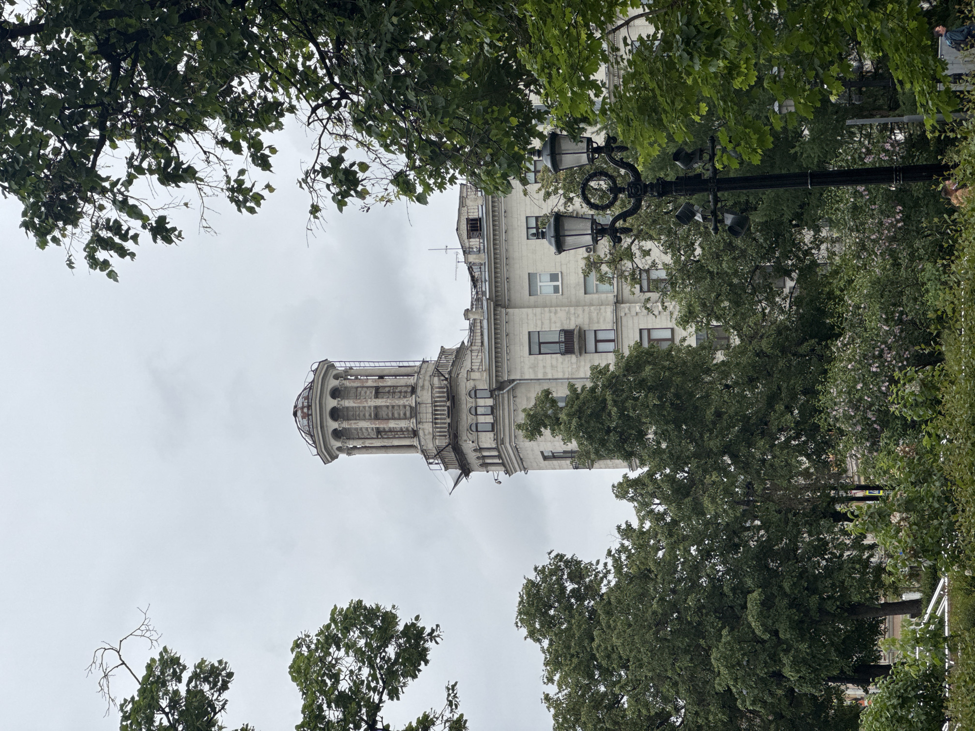 The image captures a view of the Moscow City Duma building in Moscow, Russia, framed by trees. The architectural style is a prominent feature. A streetlamp is seen to the right of the tower. The sky is overcast. The viewpoint is from below, looking up.