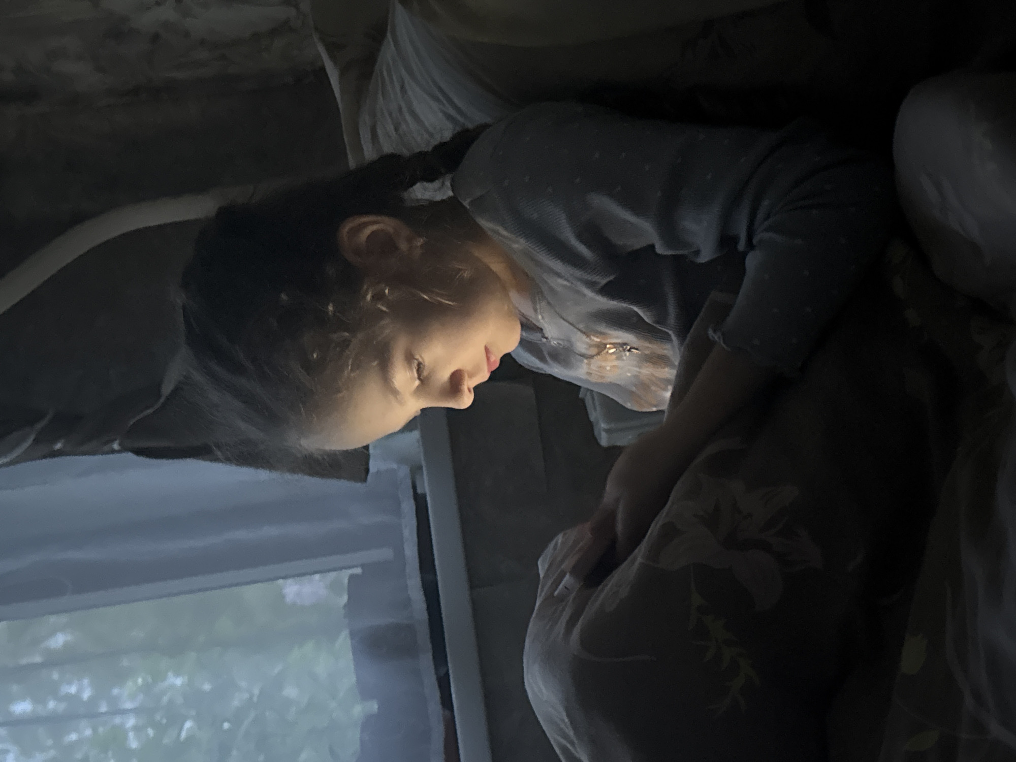 A young girl lies in bed, possibly looking at a mobile phone. She is covered in a blanket with a floral print. There is a window in the background, and a ceiling visible in the upper portion of the image. There is no evidence to link the image to the city of Раздолье, Russia.