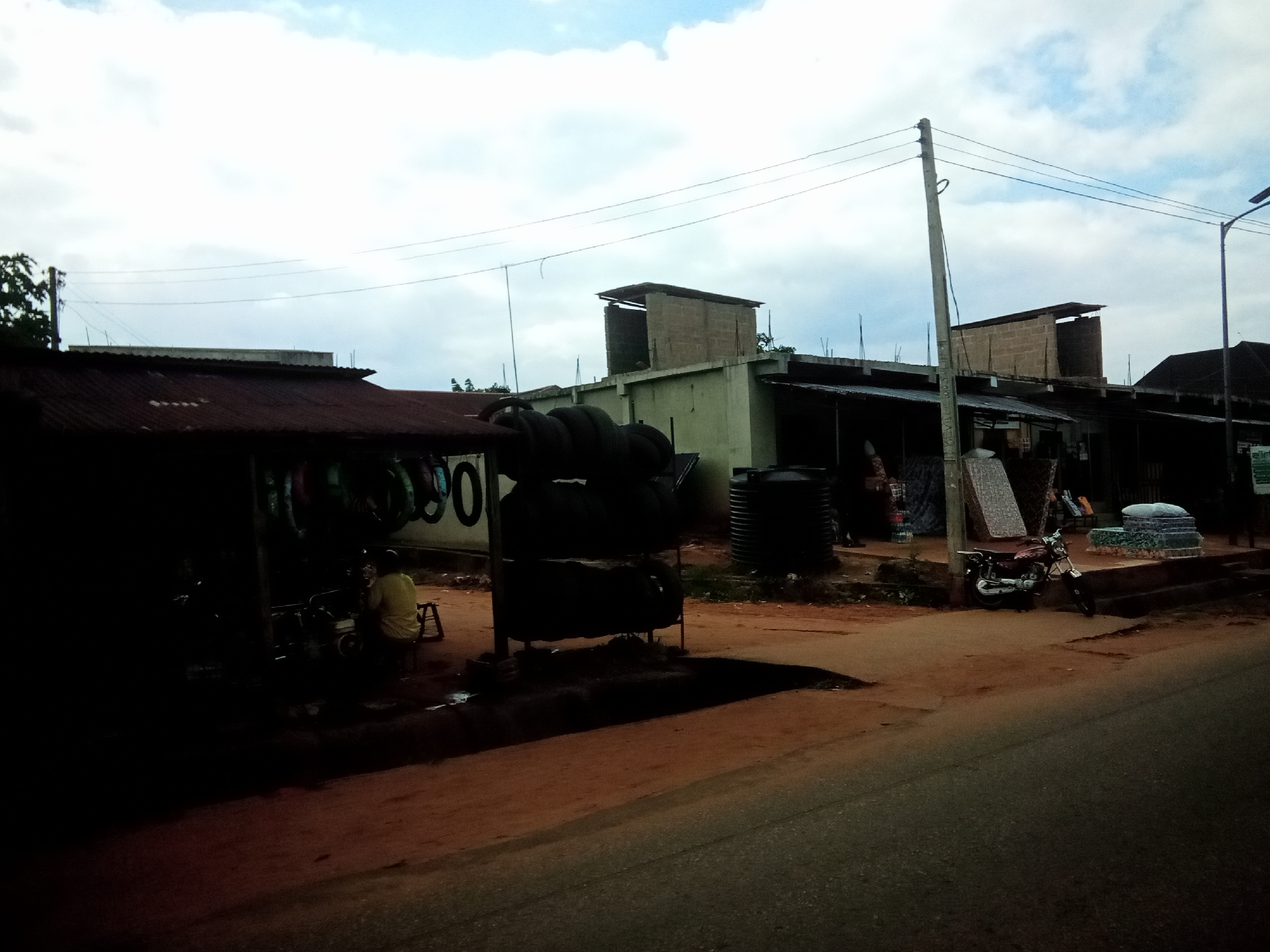 The image depicts a street scene in Nibo, Nigeria, likely in front of a commercial establishment. On the left, a covered shop displays stacks of tires, with a person in a yellow shirt visible sitting on a stool. In the background, several buildings with corrugated metal roofs and concrete structures are present. A motorcycle is parked on the right side of the street. The ground is a mixture of dirt and asphalt. Power lines crisscross the sky, which is partly cloudy. The overall impression is of a typical market or roadside business area.
