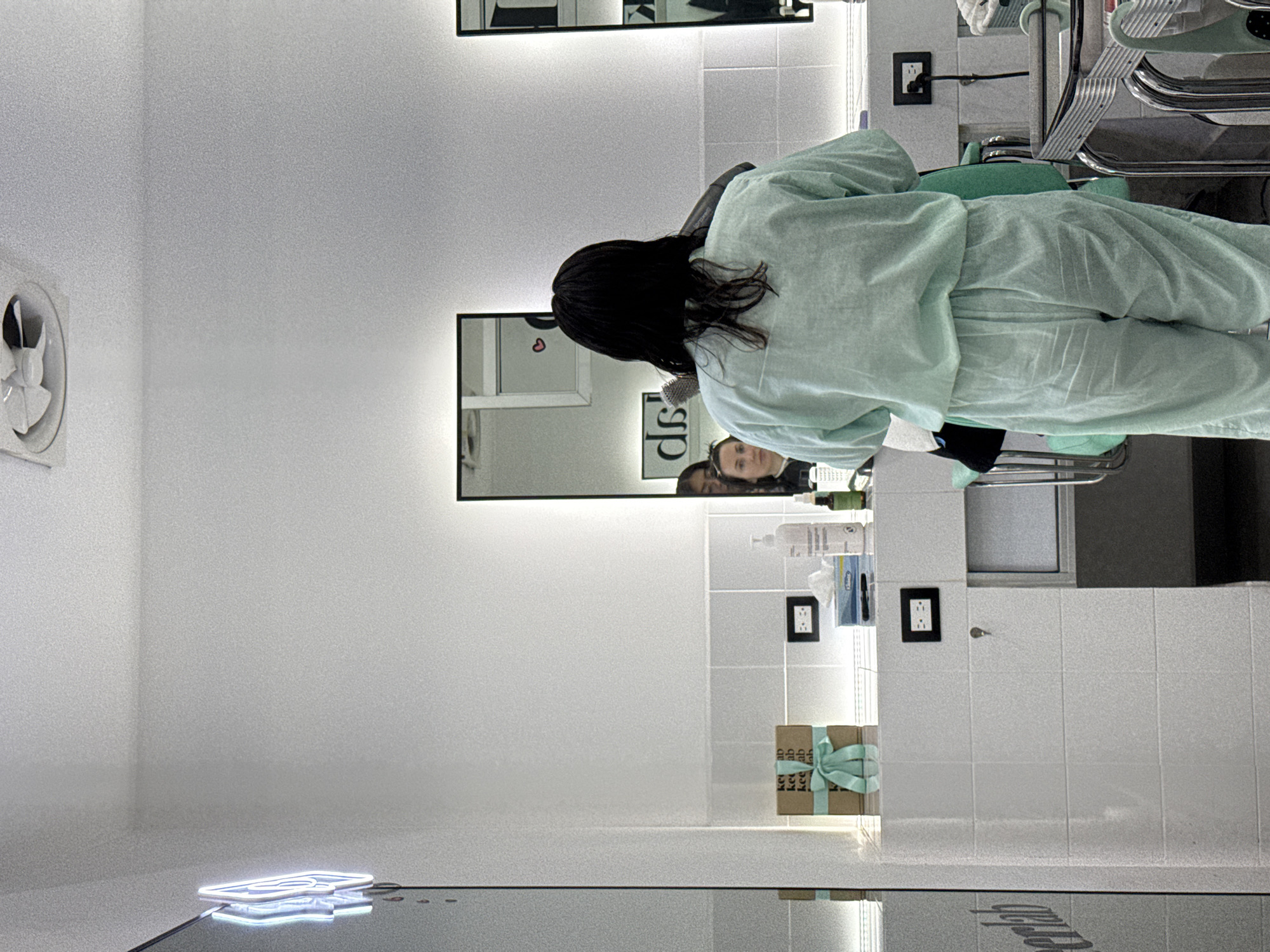 A woman with dark hair wearing a light green medical gown sits in a salon chair in a well-lit room. Two other women are visible in the mirror, both wearing dark shirts. The background shows white-tiled walls, multiple mirrors, various hair care products, and electrical outlets. The image may be taken in a salon in San Bernardino Tlaxcalancingo, Mexico.
