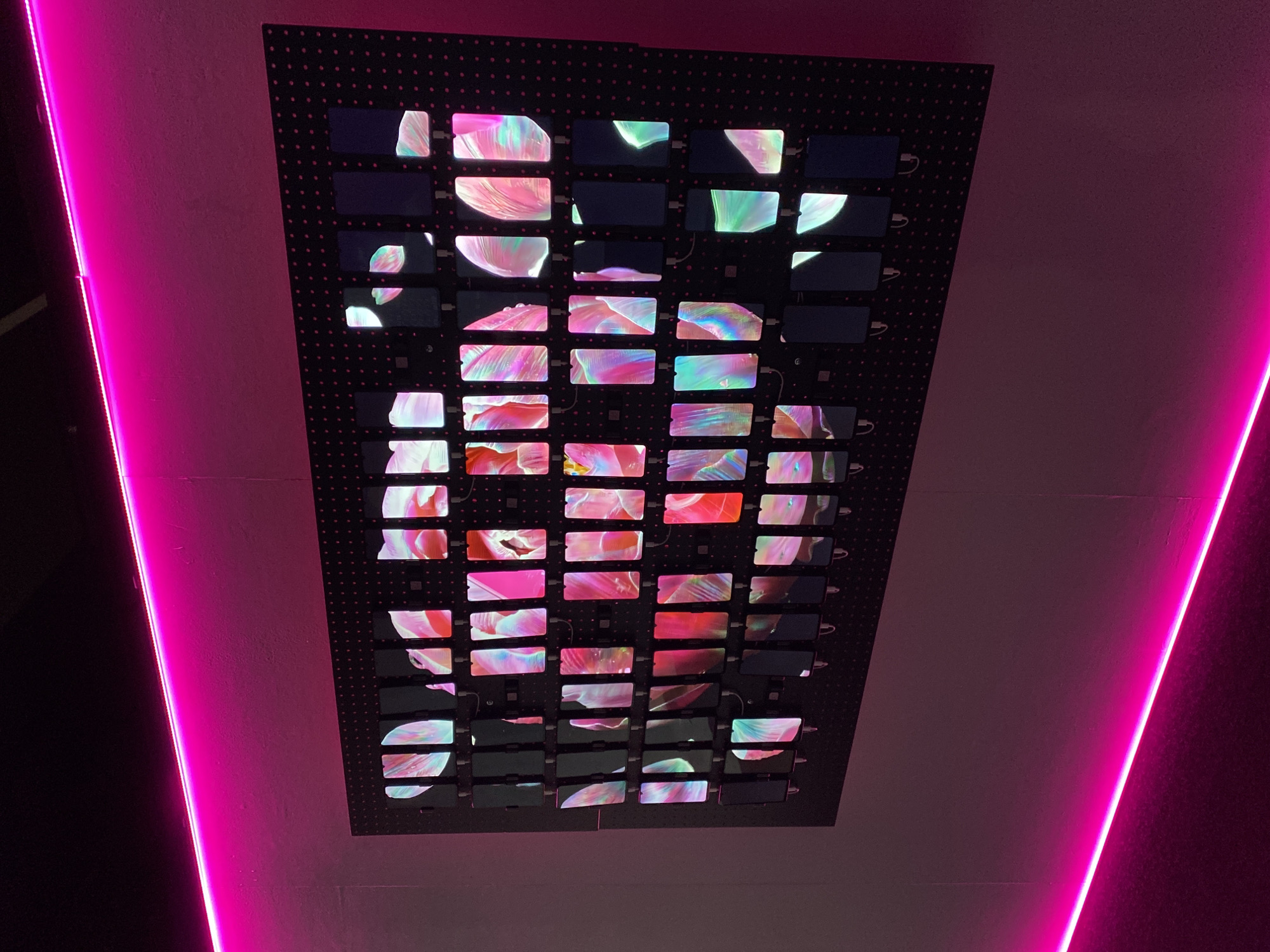 The image captures a close-up view of a modern art installation located in Yekaterinburg, Russia. The installation consists of a dark, rectangular grid-like structure mounted on a wall. Within the grid, numerous small screens display a fragmented, abstract moving image of red, pink, white, and green swirling patterns. The surrounding environment is illuminated by vivid pink neon lights positioned along the edges of the wall, creating a contrasting visual effect. The rest of the wall is a pale pink color.