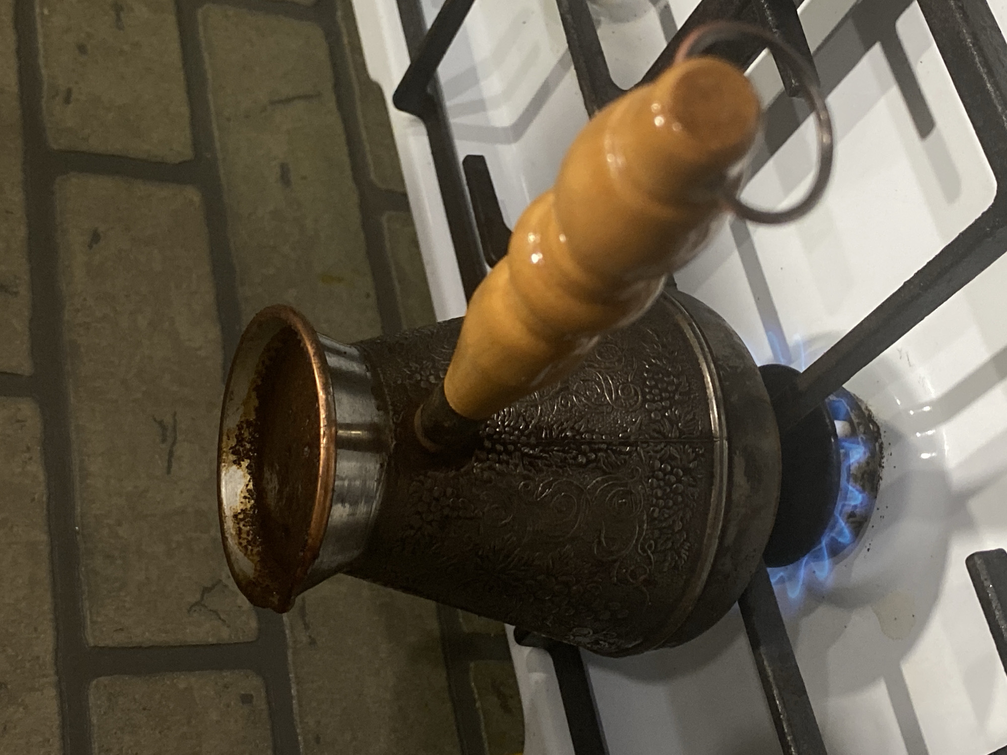A dark, ornately patterned copper or brass cezve with a flared, bright copper rim and a light brown wooden handle rests on a white gas stovetop. The cezve contains a dark brown liquid, consistent with brewing coffee, with visible grounds or foam residue around the interior upper edge. A small, vibrant blue gas flame is active beneath the cezve from a central burner on the stovetop. The stovetop is fitted with black, cast-iron grates. The background consists of a wall tiled with light brown or tan rectangular brick-like units with dark grout lines. No individuals are present. The image does not contain any visual identifiers that specify its location as Sochi, Russia; the scene depicts a generic kitchen environment.