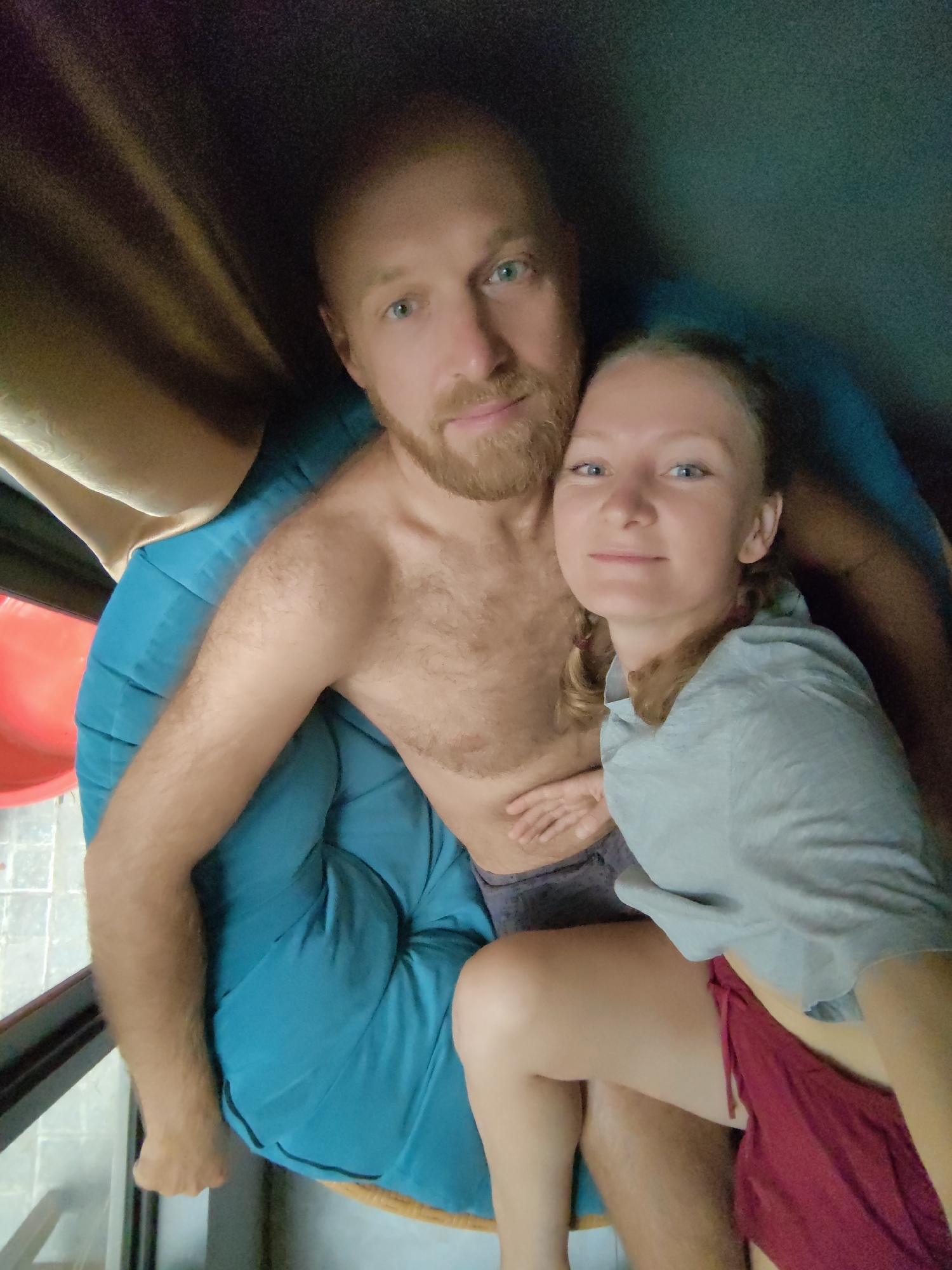 An intimate, high-angle selfie captures a man and a woman seated closely together on a large, plush teal cushion or beanbag chair. The man, positioned slightly behind and to the left of the woman, is shirtless, revealing a hairy chest. He has a reddish-blonde beard and light-colored eyes, and he looks directly at the camera with a calm expression. The woman, with blonde hair that appears to be styled in braids or pigtails, is wearing a light grey short-sleeved top and a red garment on her lower body, possibly shorts or a skirt. She leans slightly against the man and smiles gently at the camera. Their relaxed postures and close proximity suggest comfort and affection.
The setting appears to be an indoor or covered outdoor area, with beige curtains visible in the upper left background and a tiled floor in the lower left. A bright red, curved object, possibly part of a child's slide or a piece of furniture, is partially visible behind the man on the left side of the frame. The lighting is soft, creating a cozy atmosphere. No text is visible in the image. The location context provided is Hà Nội, Vietnam.