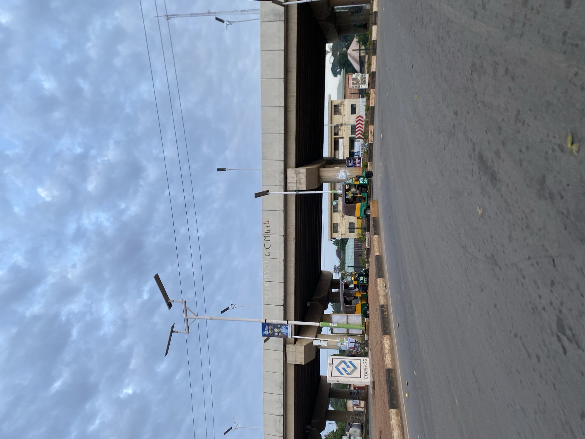 This is a medium shot, eye-level, outdoor photograph taken on a cloudy day, likely in the city of Jos, Nigeria. The foreground features a paved road with scattered leaves. Beyond the road is a raised concrete overpass with supporting pillars. Beneath the overpass, several three-wheeled vehicles, known as "Keke" or "Marwa," are parked. Two Keke are clearly visible: one is green and yellow, with license plate "PTA 829". Another Keke, predominantly yellow, is parked slightly further back and to the right. A white sign with a blue logo and the word "CRANEBURG" is positioned on the left side of the road. Several lampposts with solar panels are visible, including one to the left of the CRANEBURG sign and others beneath the overpass and along the road. There is graffiti on the underside of the overpass, including the text "MUCHIKA 27 GCM 4+4". In the background, buildings with red roofs and light-colored walls can be seen. The sky is filled with a dense, overcast layer of gray clouds.