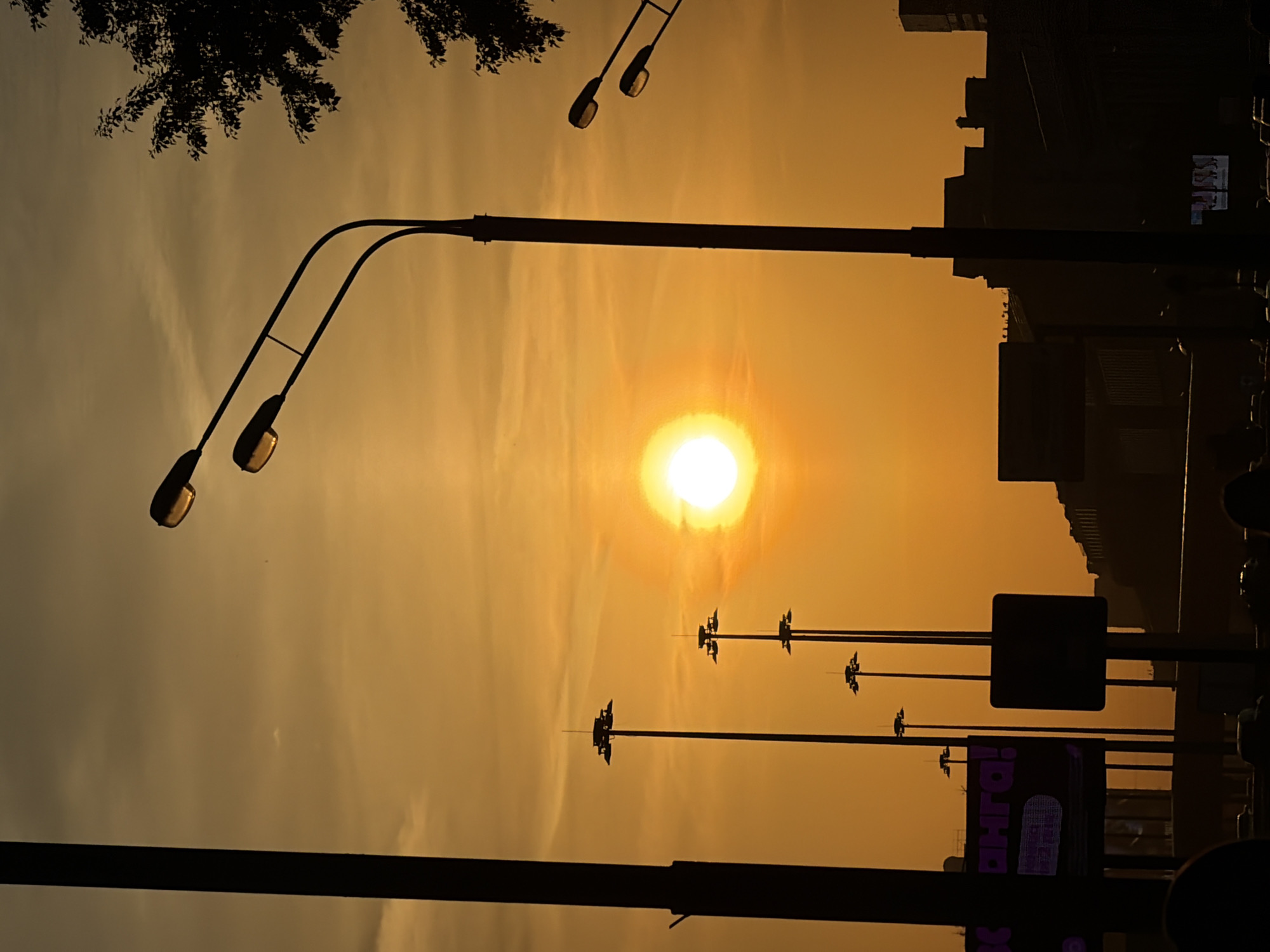The image depicts a sunset scene in a city. The sun is prominent in the sky, radiating a bright, circular glow. Silhouetted objects are scattered throughout the frame, including streetlights with curved arms, utility poles topped with equipment, buildings with varying heights, a sign, and trees with a dense cluster of leaves. The sky transitions in color from light yellow to a darker orange hue.