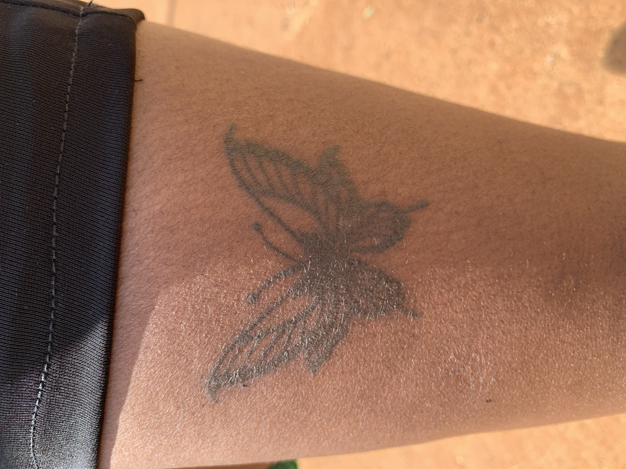 The image shows a close-up of a person's leg with a tattoo of a butterfly on it. The person is wearing black shorts. The background is blurred and appears to be a reddish-brown surface, possibly dirt or sand. There is no visible indication of the city of Jos, Nigeria.