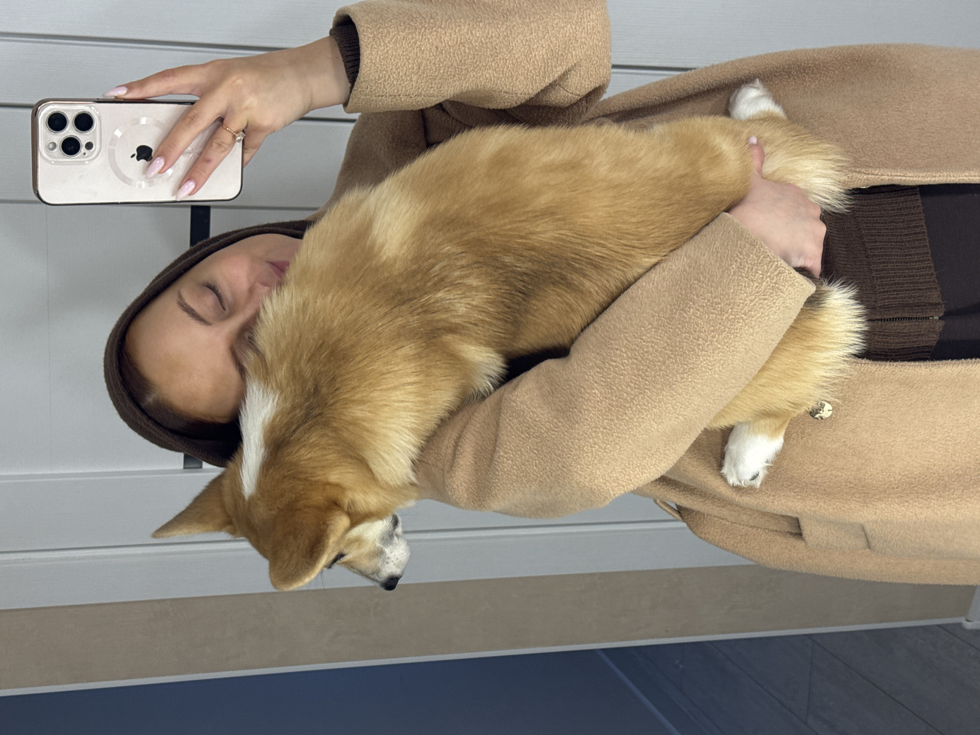 The image shows a woman taking a selfie while affectionately holding a dog indoors in Chaikovsky, Russia. The woman's face is partially visible, with her eyes closed or cast downwards, suggesting a moment of peace or connection with her pet. She is wearing a light brown or tan long coat with a textured surface, layered over a dark brown ribbed garment. Her left hand holds a white smartphone, which appears to be an iPhone with a triple camera setup and a visible MagSafe ring on its back, enclosed in a clear case. She has long, light pink manicured nails and a ring on her left ring finger.
Cradled closely to her chest is a medium-sized dog, resembling a Corgi, with golden-brown fur and distinctive white markings on its head and paws. The dog appears calm and relaxed in her arms.
The background suggests a modern indoor setting. It features white horizontal paneling, possibly a door or wall, in the upper section. Below this, there is a horizontal beige strip followed by a dark gray or blue floor, which could be wood or tile. The lighting is soft, consistent with an indoor daytime scene. The overall impression is one of warmth, comfort, and a strong bond between the woman and her dog.