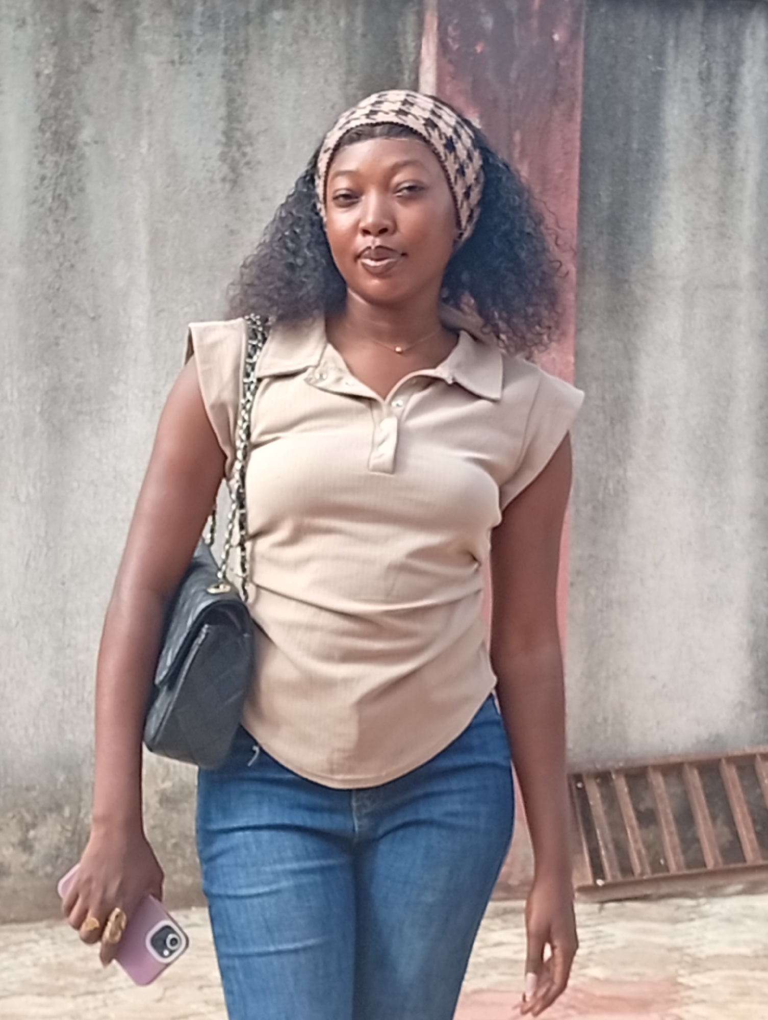 A woman stands in front of a concrete wall. She is wearing a beige polo shirt and blue jeans, with a patterned headband. A black purse hangs on her left shoulder and she holds a smartphone in her left hand. There is a metal grate on the ground near her. The location is Unknown, Unknown.