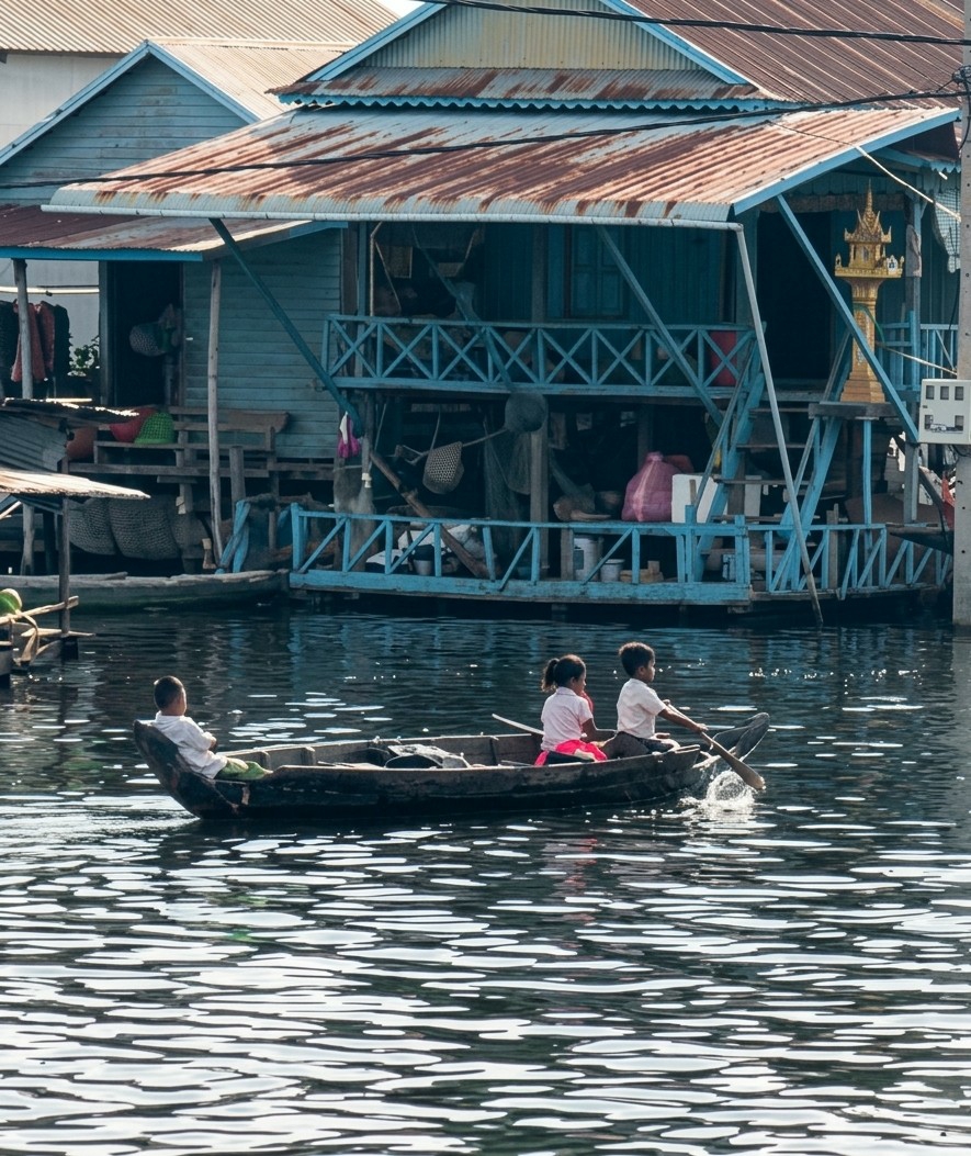 In the city of Soutr Nikom, Cambodia, a boat carries three children on a body of water. The boat is being rowed by a boy in the rear, who is wearing a white shirt and dark pants. Beside him, a girl in a pink shirt and pink skirt sits facing forward. In the front of the boat, a boy in a white shirt and green pants sits facing backward. Behind the boat, a stilted structure, possibly a house or market, is built over the water. The structure has a blue exterior with a rusty tin roof and multiple levels connected by stairs and railings. The water's surface is disturbed by ripples, reflecting the sunlight.