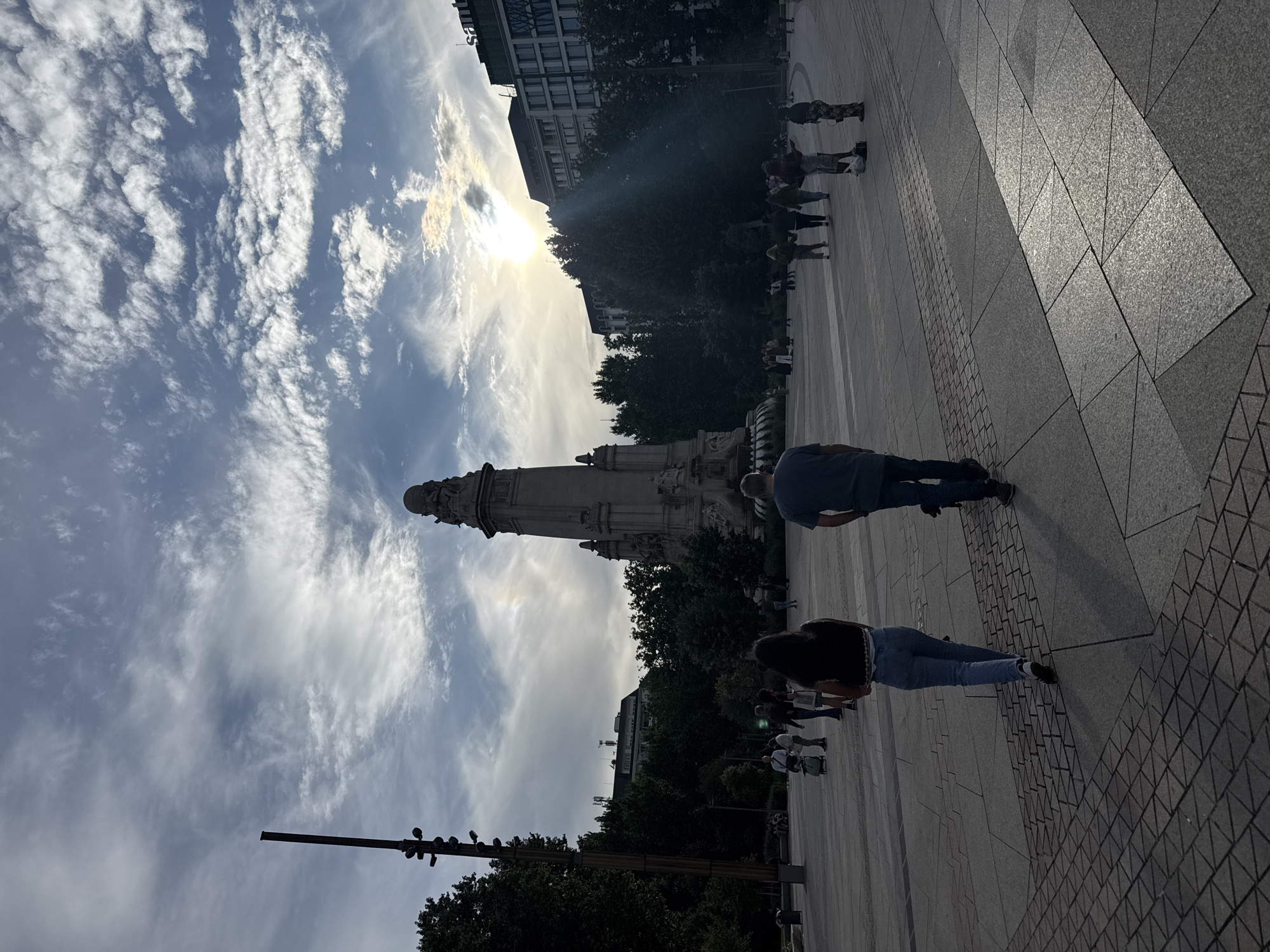 The image depicts a monument in Madrid, Spain, under a cloudy sky. The monument is tall and ornate, with multiple tiers and sculptures. Several people are walking around the plaza where the monument stands. The plaza is paved with gray stone tiles in various patterns. Trees line the background. The sun shines brightly through the clouds, creating a halo effect around the monument.
