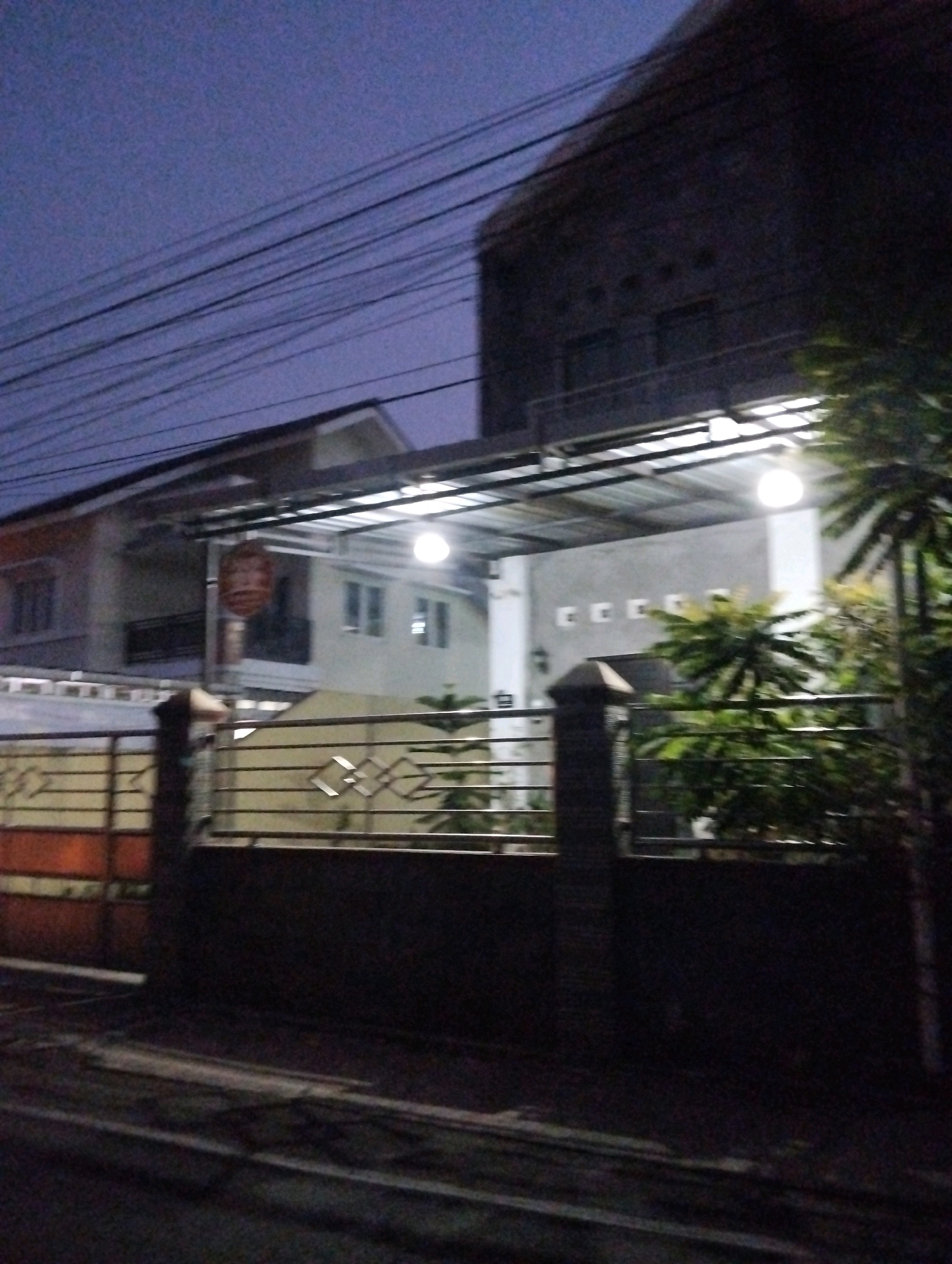 The image depicts a residential property at dusk, possibly located in Salatiga, Indonesia. The property features a two-story house with a light-colored facade and dark windows. In front of the house, a covered patio area is illuminated by several lights. A metal gate with horizontal bars and decorative elements spans the front of the property. Several power lines are visible crisscrossing the sky above the house. The overall scene suggests a quiet, residential street with moderate lighting. The image quality is somewhat grainy, likely due to low light conditions.
