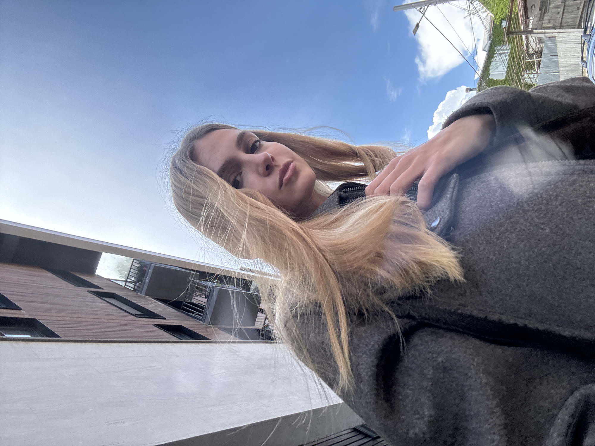A woman with long blonde hair, wearing a gray coat, stands outdoors under a blue sky with scattered clouds. The camera is angled upwards, capturing her face and upper body. A building with wooden paneling and windows is visible in the background. A power line and possibly greenery can be seen above and behind her. The location is the city of Unknown, Unknown.