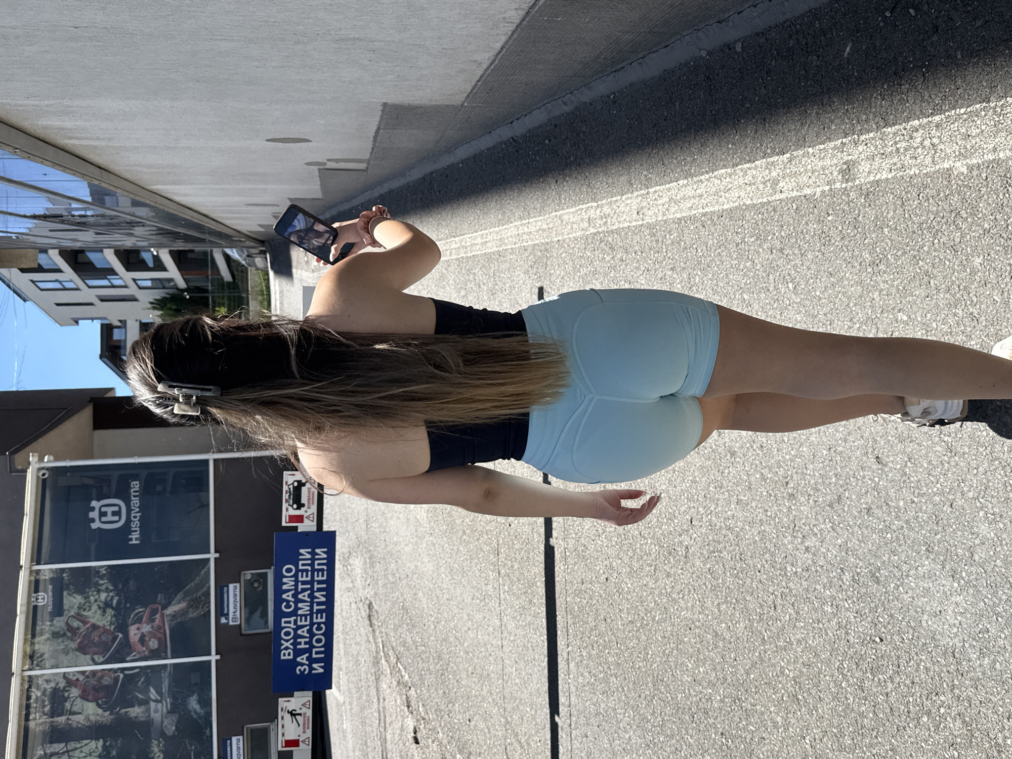 The image captures a woman from behind, standing on an asphalt road in Sofia, Bulgaria. She is holding a smartphone and appears to be taking a picture or video. She has long, brown hair and is wearing a black tank top and light blue shorts, along with white sneakers. In the background, there's a building with a Husqvarna advertisement banner, which features the brand name and images of chainsaws, along with Bulgarian text: "ВХОД САМО ЗА НАЕМАТЕЛИ И ПОСЕТИТЕЛИ," meaning "Entrance only for tenants and visitors." There's also a sign that indicates "Husqvarna" and "PROMRACTO." The surrounding environment includes buildings and the sky is visible above.