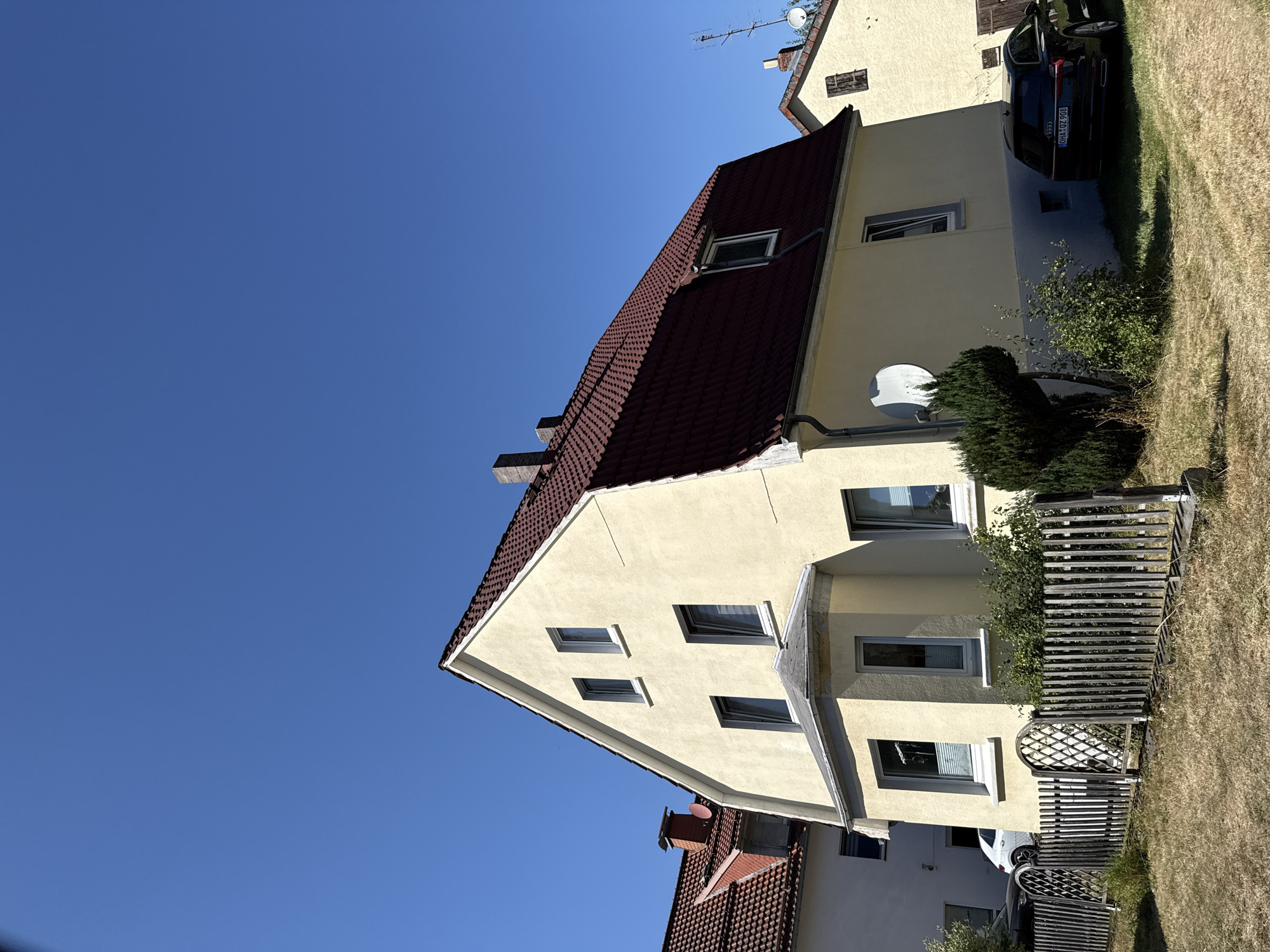 This is a medium-shot, eye-level, outdoor photograph of a residential area in Bad Lauterberg im Harz, Germany. The image is dominated by a two-story, light yellow house with a dark red tiled roof. The house has several windows, some of which are open, and a bay window on the first floor. There is a small, decorative wooden fence in the foreground, separating the property from a dry, grassy yard. A dark colored Audi station wagon is parked in the driveway on the right side of the house, with a license plate visible. To the left of the house, another lighter colored car is partially visible. The sky is clear and bright blue. Another house, also with a red tiled roof, is visible to the left of the main subject. A satellite dish is mounted on the side of the main house. The overall impression is of a sunny day in a suburban neighborhood.