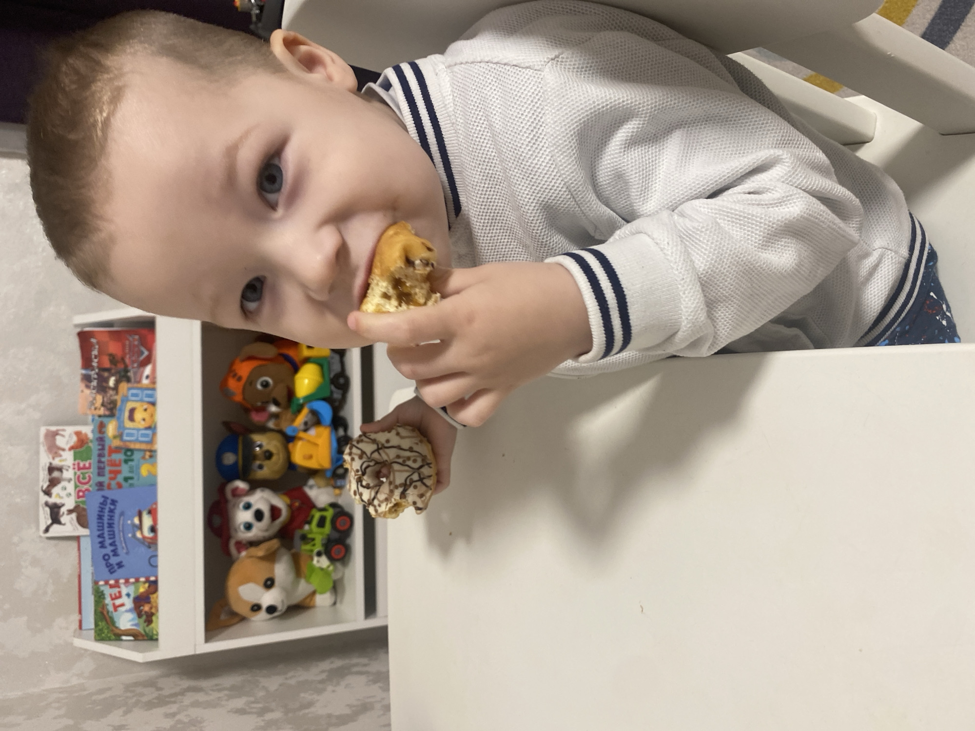 A young male child with light brown hair and blue eyes is seated at a white table, consuming food in an indoor setting. He wears a white, textured, long-sleeved shirt with navy blue stripes on the collar and cuffs. His right hand holds a partially eaten donut to his mouth, while his left hand holds a second donut with white frosting and dark drizzle patterns.
Behind the child is a white, two-shelf unit. The top shelf contains several children's books with titles such as "ПРО МАШИНКИ И МАШИНКИ" (About Cars and Cars) and "Я ПЕРВЫЙ СЧЁТ" (My First Count). The bottom shelf displays four plush toys, resembling characters from Paw Patrol (including Marshall, Chase, and Rocky), and two small toy vehicles (a green excavator and a yellow dump truck). The wall behind the shelving unit is covered with light-colored, subtly patterned wallpaper. A corner of a striped rug (yellow, blue, white) is visible on the floor to the bottom right.
The specific address or precise location within the city of Sochi, Russia, cannot be determined from the provided image. The presence of Russian language books indicates the general region.