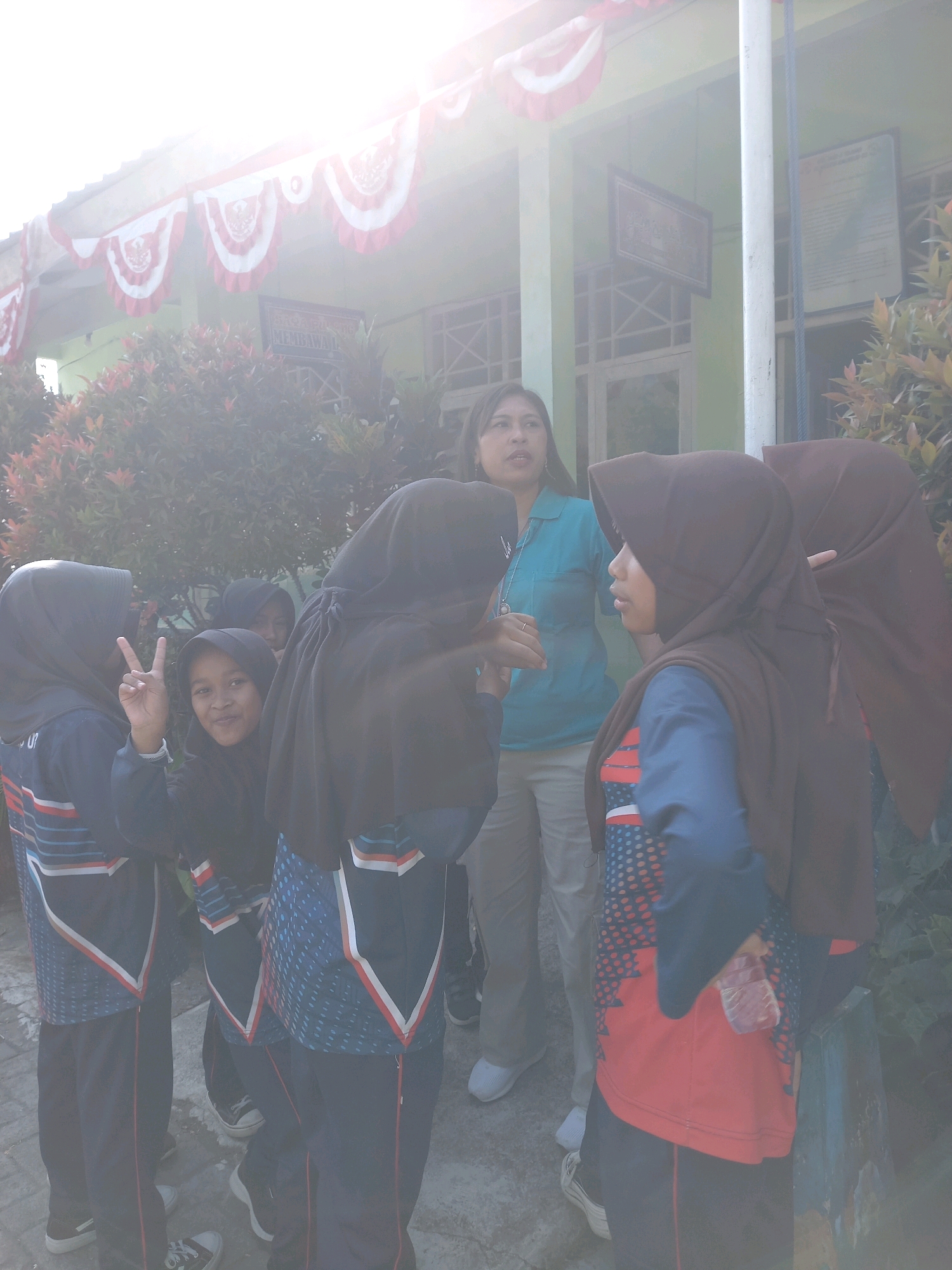 In Kebaturan, Indonesia, outside a green building adorned with red and white festive banners, a group of individuals, primarily young girls wearing hijabs and school uniforms, are gathered. The girls' uniforms consist of dark blue trousers and shirts with red and white stripe accents. Some girls are interacting, with one making a peace sign. A woman in a turquoise shirt and light-colored pants stands among them. The environment includes dense foliage and a bright, hazy sky, suggesting daytime conditions possibly impacted by sunlight glare or atmospheric conditions. The building has windows and framed pictures or signs on its exterior.