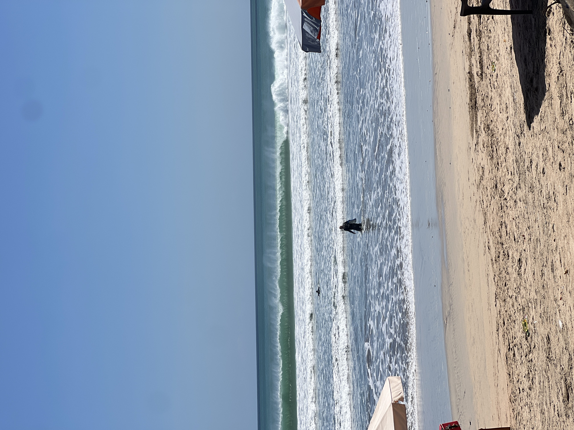 A wide shot captures a beach in Kuta, Indonesia, under a clear blue sky. Large, greenish-blue waves crash in the background, creating a frothy white surf. A person wearing a dark wetsuit walks through the shallow water towards the ocean, carrying a surfboard. Another surfer is visible in the distance, paddling in the water. In the foreground, two beach umbrellas, one beige and one orange and grey, provide shade on the sandy shore. The sand is light brown and textured with footprints and debris. A dark shadow cast by an unseen object falls across the sand in the bottom right corner.