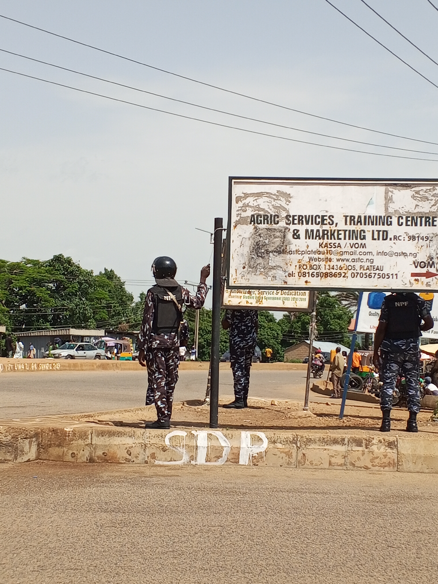 The image depicts a street scene in Bukuru, Nigeria, under a bright, overcast sky, suggesting midday. Three individuals in camouflage uniforms, likely law enforcement, are present. One individual, in the foreground on the right, stands facing away from the camera, his uniform clearly marked with "NPF" on his vest. Another individual, in the center, is also facing away and appears to be in mid-action, possibly gesturing or interacting with something out of frame. The third individual is partially visible on the left.
A prominent billboard with black text on a white background dominates the upper half of the image. The text advertises "AGRIC SERVICES, TRAINING CENTRE & MARKETING LTD." and includes contact information such as email addresses, a website, a P.O. box, and phone numbers. Below this, smaller text reads "Knowledge, Service & Dedication Courtesy Student Or Caverament (ISU) 2018/2019".
The setting appears to be a roadside intersection or busy thoroughfare. Cars and a three-wheeled vehicle (Keke Napep) are visible in the background, along with trees and buildings. The curb in the foreground has white spray-painted lettering, partially obscured and worn, with visible letters including "SIDP". The overall atmosphere seems to be one of normal daytime activity with a visible security presence.
