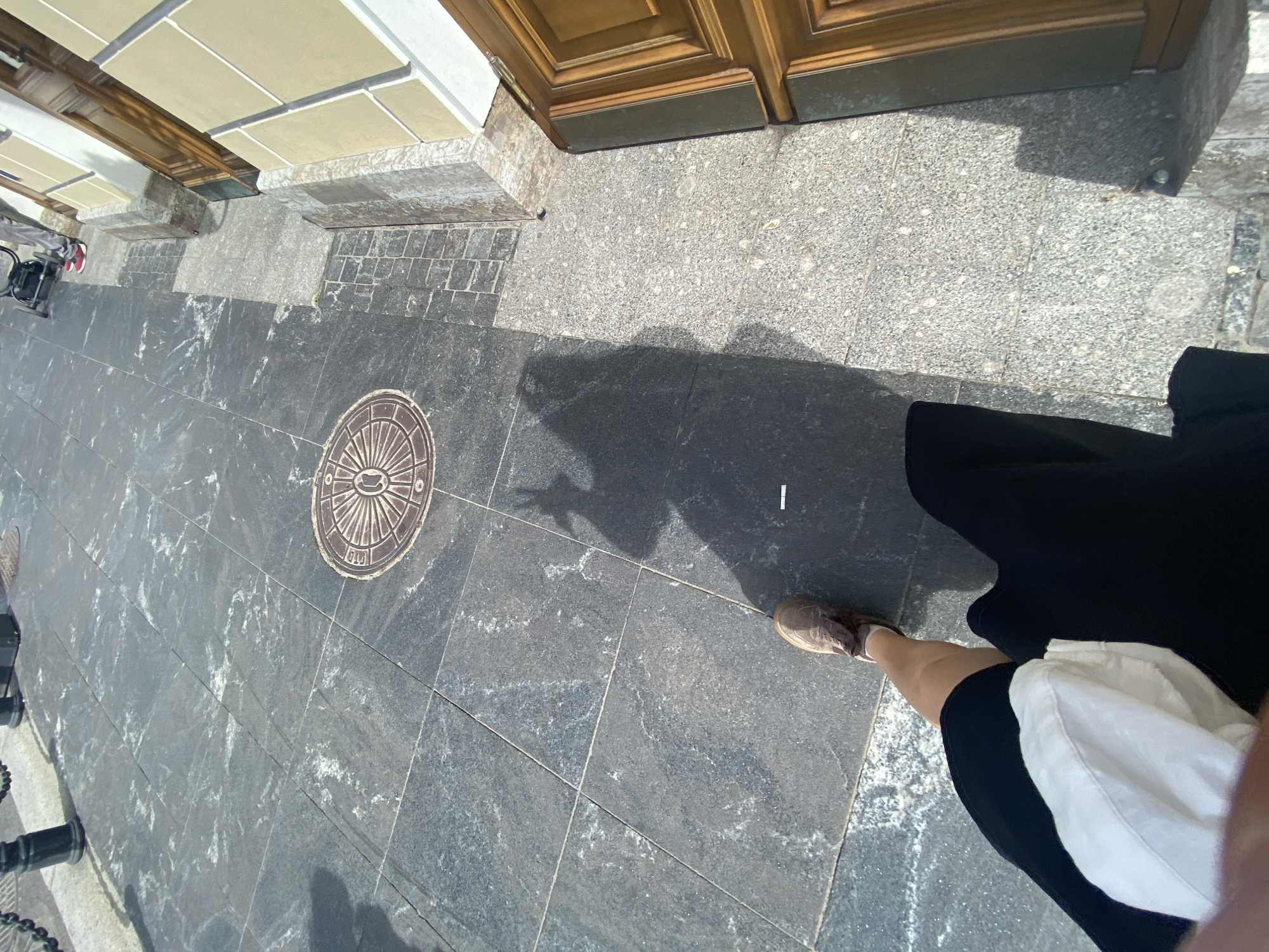 An overhead, high-angle shot shows a person's legs and shadow on a city sidewalk in Saint Petersburg, Russia. The person is wearing a black skirt and brown shoes. The sidewalk is made of large, dark granite tiles. A shadow of the person is cast on the ground, with a cigarette butt visible on the sidewalk within the shadow. In the background, a building with wooden doors and light-colored walls is visible. A circular metal manhole cover is embedded in the sidewalk. The environment suggests an urban setting on a sunny day.