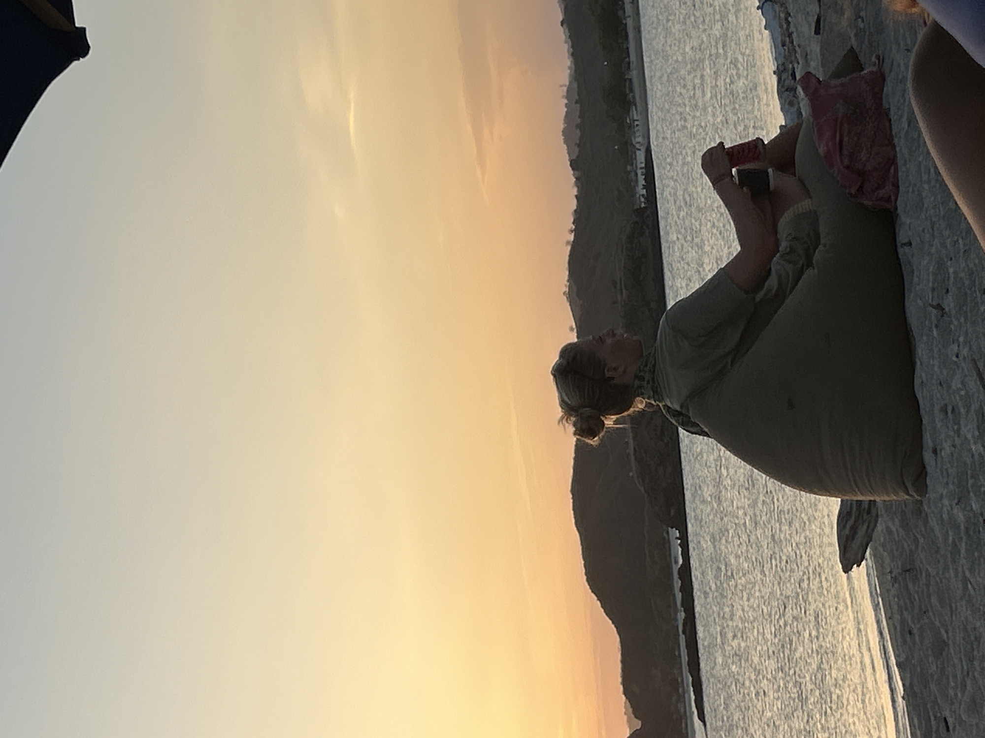 At sunset on a beach in Kuta, Indonesia, a woman with blonde hair in a bun sits in a large, beanbag-like chair on the sand. She is wearing a green shirt and is holding a red can in her right hand, appearing to be drinking from it. Beside her, a pink bag is placed on the sand. The ocean stretches out before her, with a gentle shore and distant hills silhouetted against the orange and yellow sky. A dark umbrella is partially visible in the upper right corner.