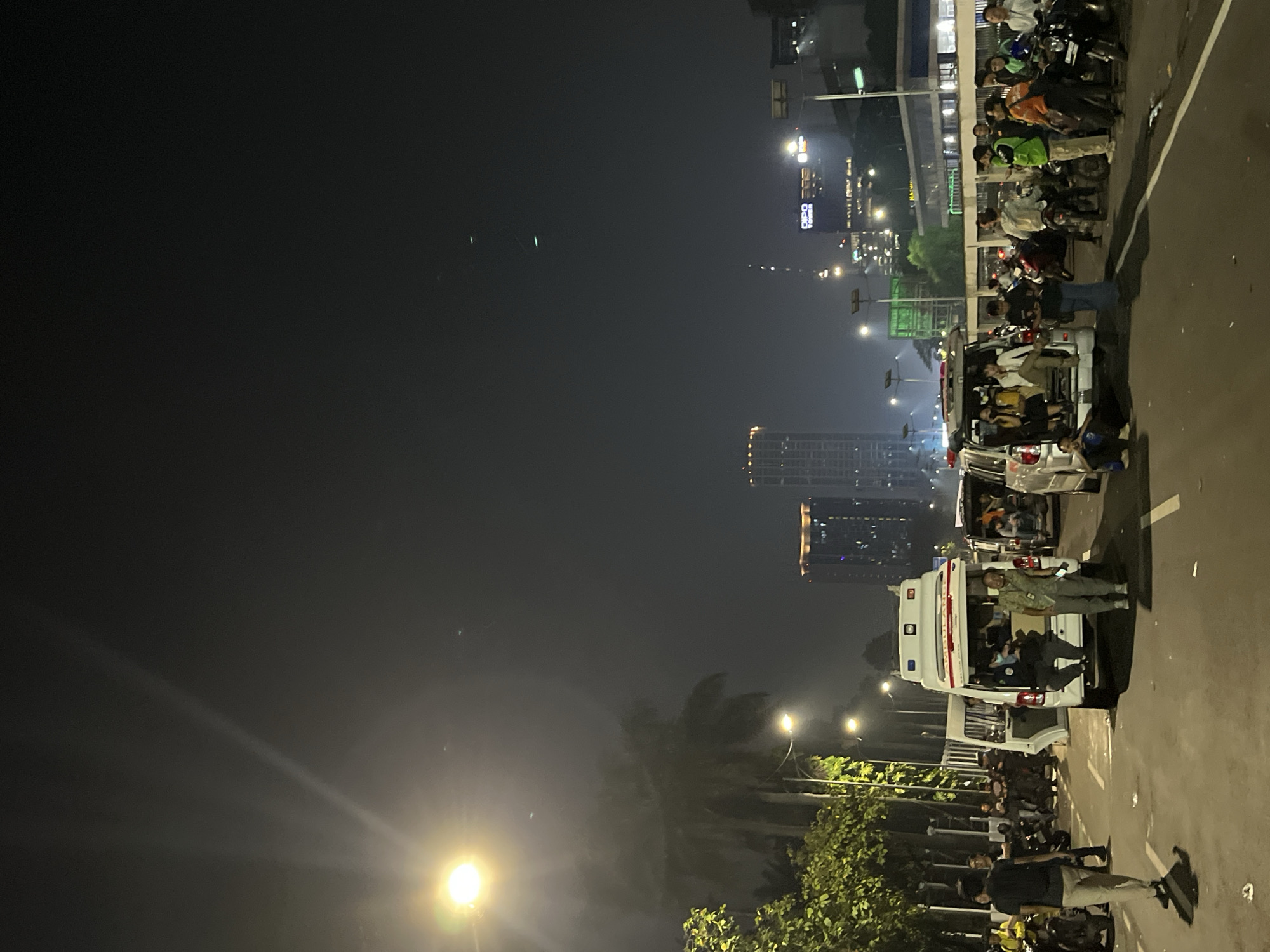 The image depicts a nighttime scene on a street in Central Jakarta, Indonesia. The overall lighting suggests it is after dark, with artificial lights from buildings, street lamps, and vehicle headlights illuminating the area. The weather appears to be clear, with no obvious signs of rain or fog, though there might be some atmospheric haze.
The primary subjects are a group of people and several vehicles, predominantly motorbikes and white vans, some of which appear to be ambulances. The people are gathered around the vehicles, with some standing and others seated or leaning on the motorbikes. One ambulance's rear doors are open, revealing people inside. Another van, also white, is parked behind the ambulance. A line of motorbikes, with riders and passengers, stretches along the side of the road.
The activity appears to be a gathering or a temporary stop, possibly related to an event or an emergency situation, given the presence of what look like ambulances. People are casually dressed, and their postures don't suggest an immediate sense of panic or distress, but rather a state of waiting or observation. The background features urban elements: tall buildings, some illuminated, indicating a city environment. One building displays the text "DIPO TOWER" in large letters, providing a specific location cue. The street itself is paved, and there's a clear demarcation of a lane. The presence of trees and greenery suggests a street lined with some vegetation. The overall atmosphere is one of urban nightlife with a noticeable congregation of people and vehicles.
