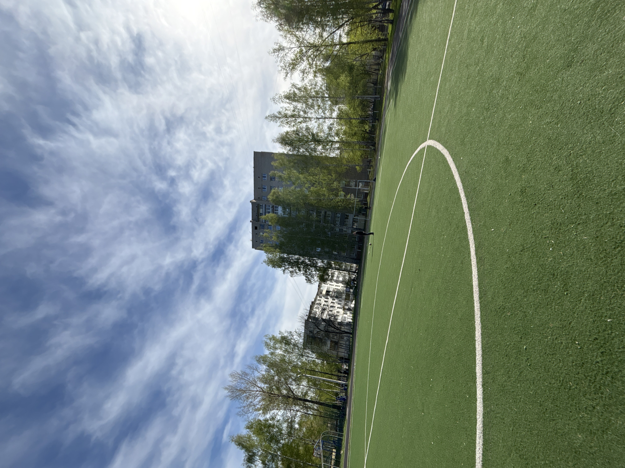 The image depicts an outdoor sports field, likely a soccer field, featuring artificial green turf and white line markings, including part of the center circle. The field is adjacent to a residential area. In the background, there are a few gray, multi-story buildings and a row of deciduous trees with budding green leaves. The sky is partly cloudy with a mix of blue and white. It is likely located in the city of Chaikovsky, Russia. No people are clearly visible in the scene. The overall impression is that of a public park or recreational area in an urban setting.