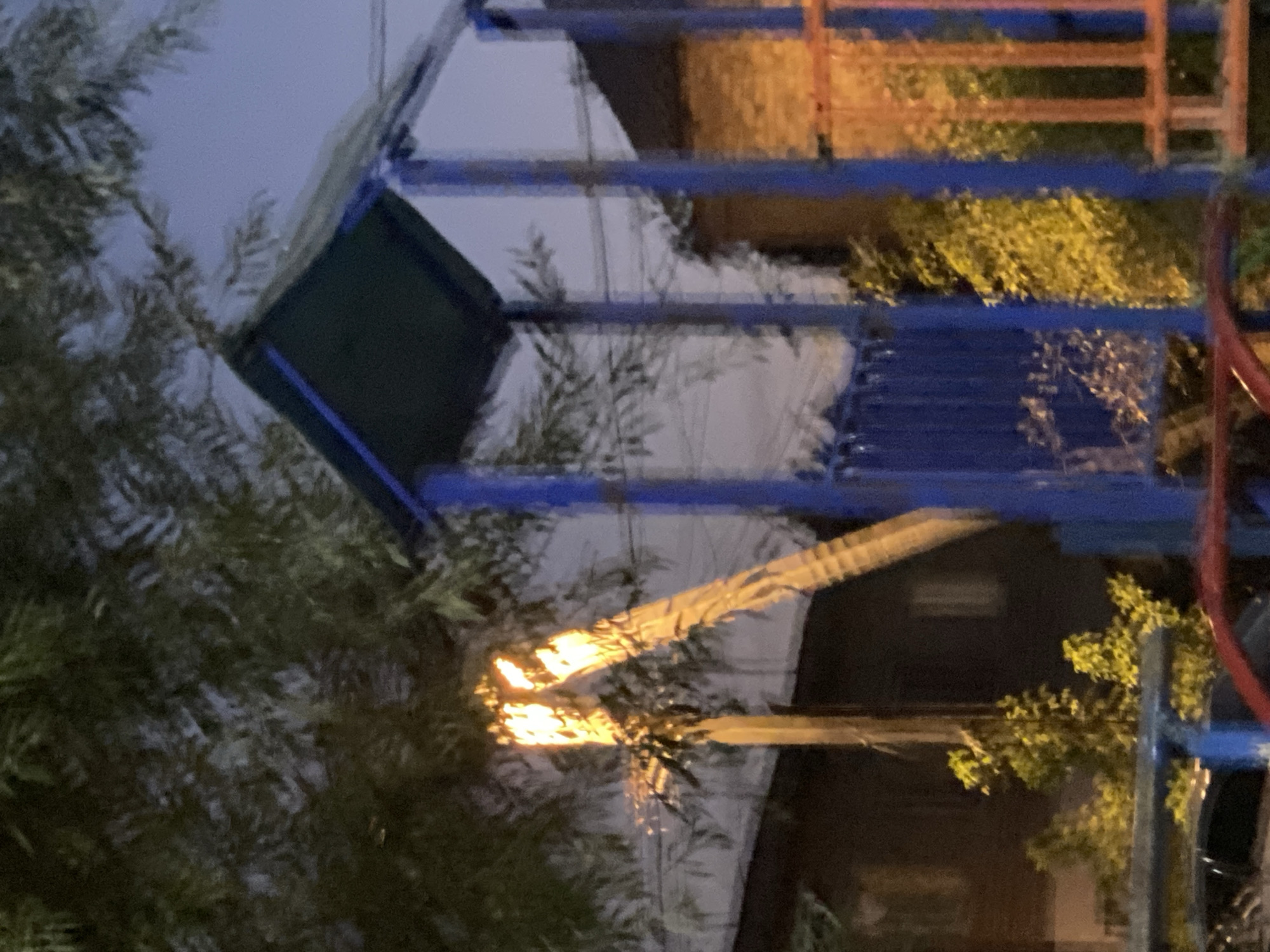 The image shows an outdoor scene at dusk or night, likely in Novopavlovka, Kyrgyzstan. In the foreground, a blue and green playground structure is visible, with blue poles and a green roof. To the left, branches of a tree are illuminated by a bright yellow light source, possibly a lamp or floodlight, which casts a strong glow on the trunk and surrounding foliage. The light appears to be concentrated on the upper part of a vertical pole or tree trunk. Behind the playground structure, buildings with brick and wood siding are dimly visible. The sky is a pale blue, indicating twilight. There are no people visible in the image. The overall impression is of a playground area illuminated at night, with a focus on a bright light source within the trees.