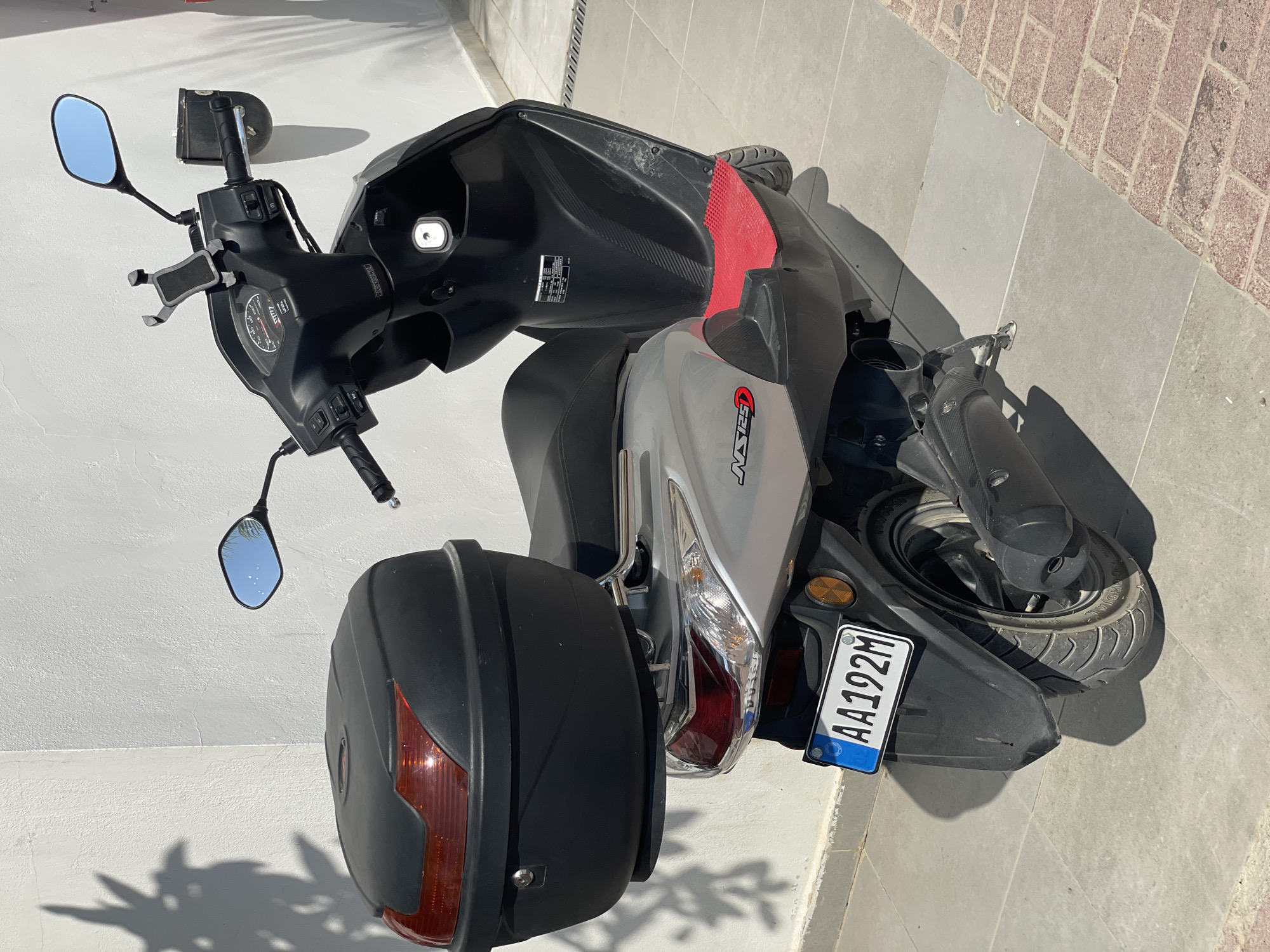 A silver and black scooter is parked on a tiled walkway in front of a white stucco wall. The scooter has a black storage box mounted on the rear, and a license plate with the characters "AA192M". The scooter's dashboard and handlebars are visible, along with a phone mount. Shadows of palm fronds are cast on the wall and the ground. The scene appears to be in a city, likely Trikomo, Northern Cyprus / Cyprus, given the European style license plate.
