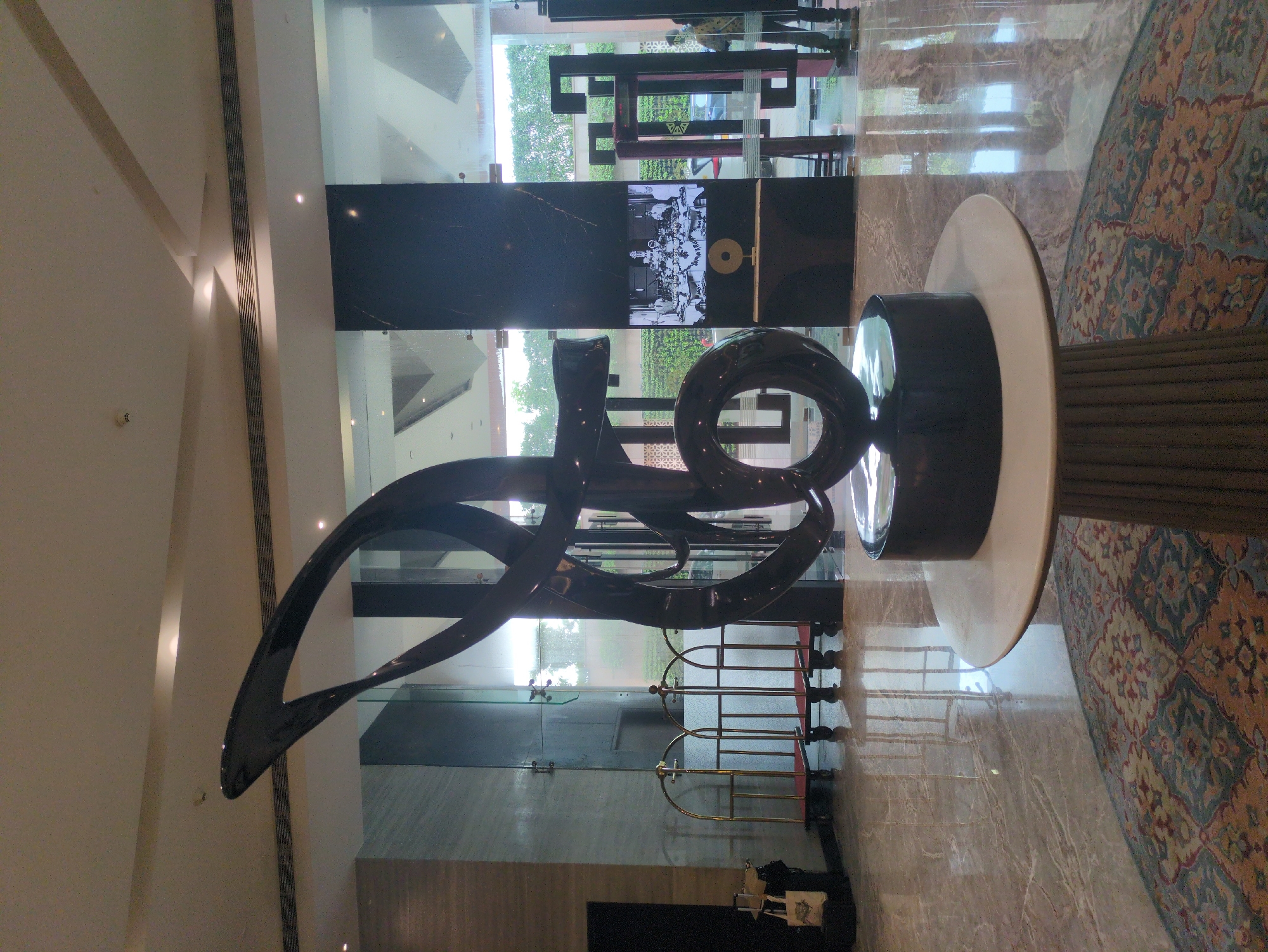 The image shows the lobby of a hotel in Agra, India. In the foreground, a large, abstract black sculpture stands on a round white pedestal, which is supported by a fluted base. The sculpture has smooth, flowing curves and appears to be made of a polished material. The lobby floor is made of polished marble with a patterned rug in front of the sculpture. Behind the sculpture, there are floor-to-ceiling glass windows that offer a view of greenery outside. To the left of the sculpture, there are several gold luggage trolleys. To the right, a digital screen displays a black and white image of a group of people around a table. The ceiling is modern, with recessed lighting and ventilation systems. The overall impression is one of a well-maintained, contemporary hotel lobby.