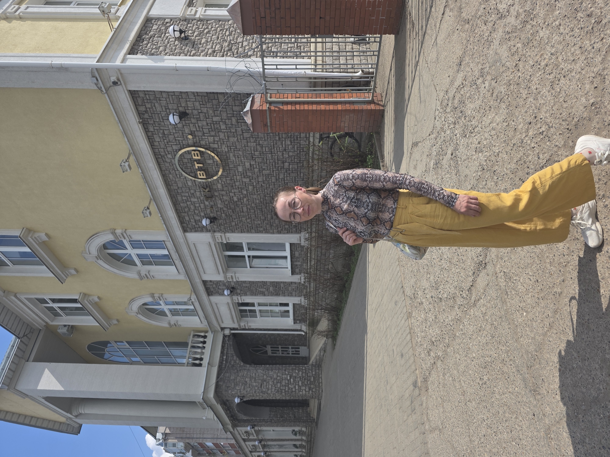 A young woman stands outside the building that houses the VTB bank branch in Votkinsk, Russia. The bank is a multi-story structure with a mix of yellow and stone facades, and arched windows. The woman is wearing a snake print top, yellow pants, and white sneakers. She stands in front of the building, slightly turned towards the camera. The ground is a mix of concrete and brick pavement.