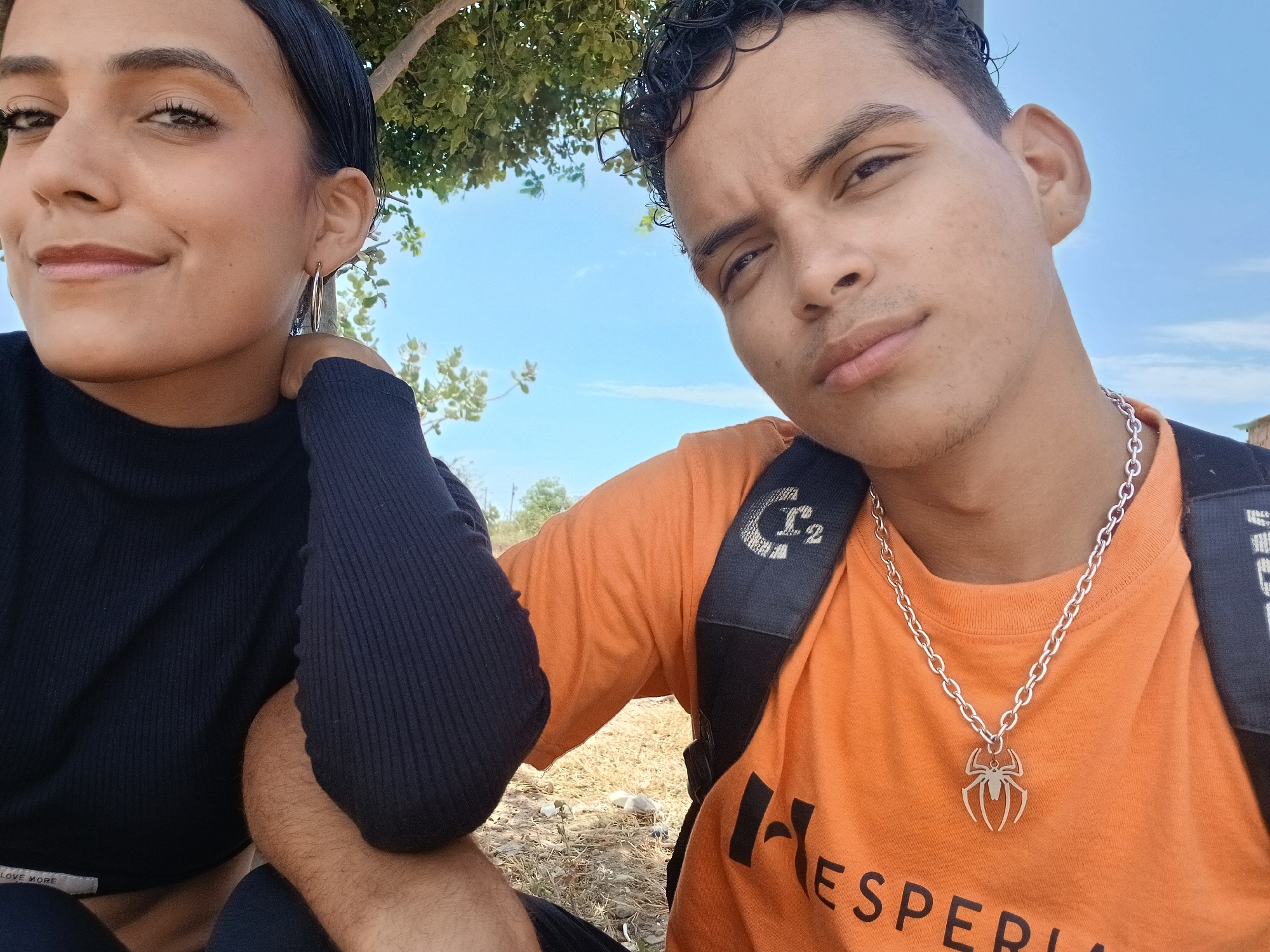 The image displays two young individuals, a female and a male, posing for a close-up outdoor photo in a bright, natural setting. The scene is consistent with a sunny day in Altagracia, Venezuela.
The female, on the left side of the frame, has dark hair pulled back and looks slightly past the camera with a gentle, relaxed smile. She wears a black ribbed long-sleeved top, on which the text "LOVE MORE" is partially visible at the bottom. She also wears large gold hoop earrings. Her right arm is bent, with her hand near her ear.
The male, on the right, has dark, curly hair and looks directly at the camera with a neutral to slightly serious expression. He is wearing an orange short-sleeved t-shirt, with the word "HESPERIA" visible on the lower left portion. Around his neck, he sports a silver chain necklace with a prominent spider-shaped pendant. A black backpack strap, featuring a stylized "C2" logo and partially visible text "MOM," crosses his right shoulder. His left arm is bent, positioned between the two subjects.
The background features a clear, bright blue sky with a few wispy clouds and vibrant green tree foliage, suggesting strong daylight, likely midday or early afternoon. The ground beneath them is dry earth with sparse vegetation, indicative of an outdoor, possibly rural or semi-rural, environment.