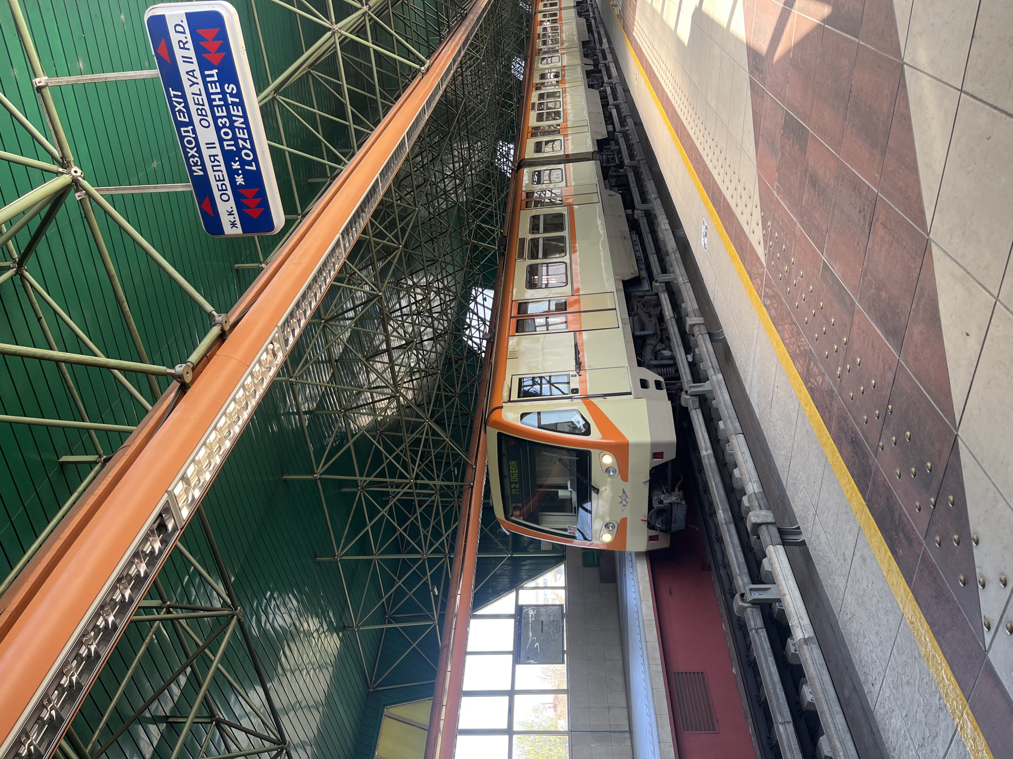 A metro train at a station. The train is cream and orange. The platform is marked with a yellow line and tiled with light and dark tiles. An overhead sign indicates the direction as "Exit", "Obelya II", and "Lozenets". This indicates the station is in Sofia, Bulgaria. The station roof is green and supported by a metal structure.