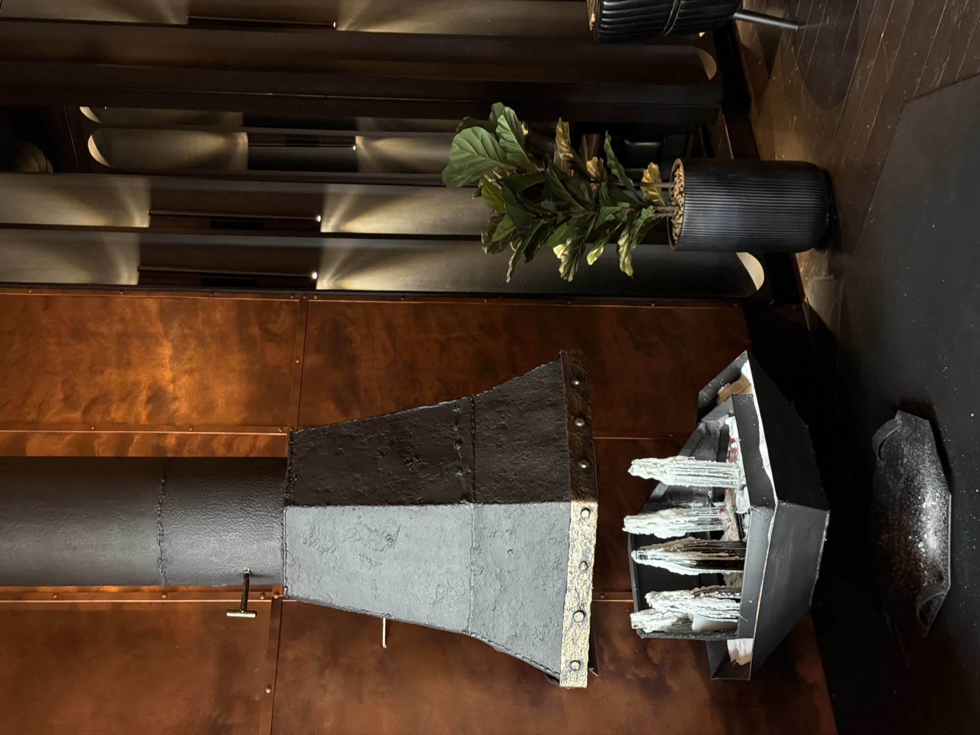 The image captures an indoor scene, likely an upscale restaurant or lounge, potentially located in Moscow, Russia, given the style of decor and the presence of luxury design elements. The setting is characterized by a mix of industrial and modern aesthetics.
The background consists of horizontally stacked, metallic-looking panels, possibly part of a wall or room divider. These panels have embedded lighting that creates a luminous effect, adding to the ambience. A potted plant with lush green leaves is placed along the top of these panels.
The foreground prominently features a copper-colored wall or structure. A dark, industrial-looking pipe or duct extends horizontally across the wall, possibly part of a ventilation or heating system. Attached to the pipe is a cone-shaped element, textured with metallic hardware details.
On the right side, there is a three dimensional sculpture, resembling a cluster of white crystalline structures, possibly made of plaster or a similar material, encased in a dark, geometric frame. A separate, smaller object made of a similar material sits to the right of the sculpture.
The floor is dark and appears to be made of wood or a similar material.
Overall, the scene portrays a well-designed and visually interesting interior space with a focus on texture, lighting, and unique decorative elements.