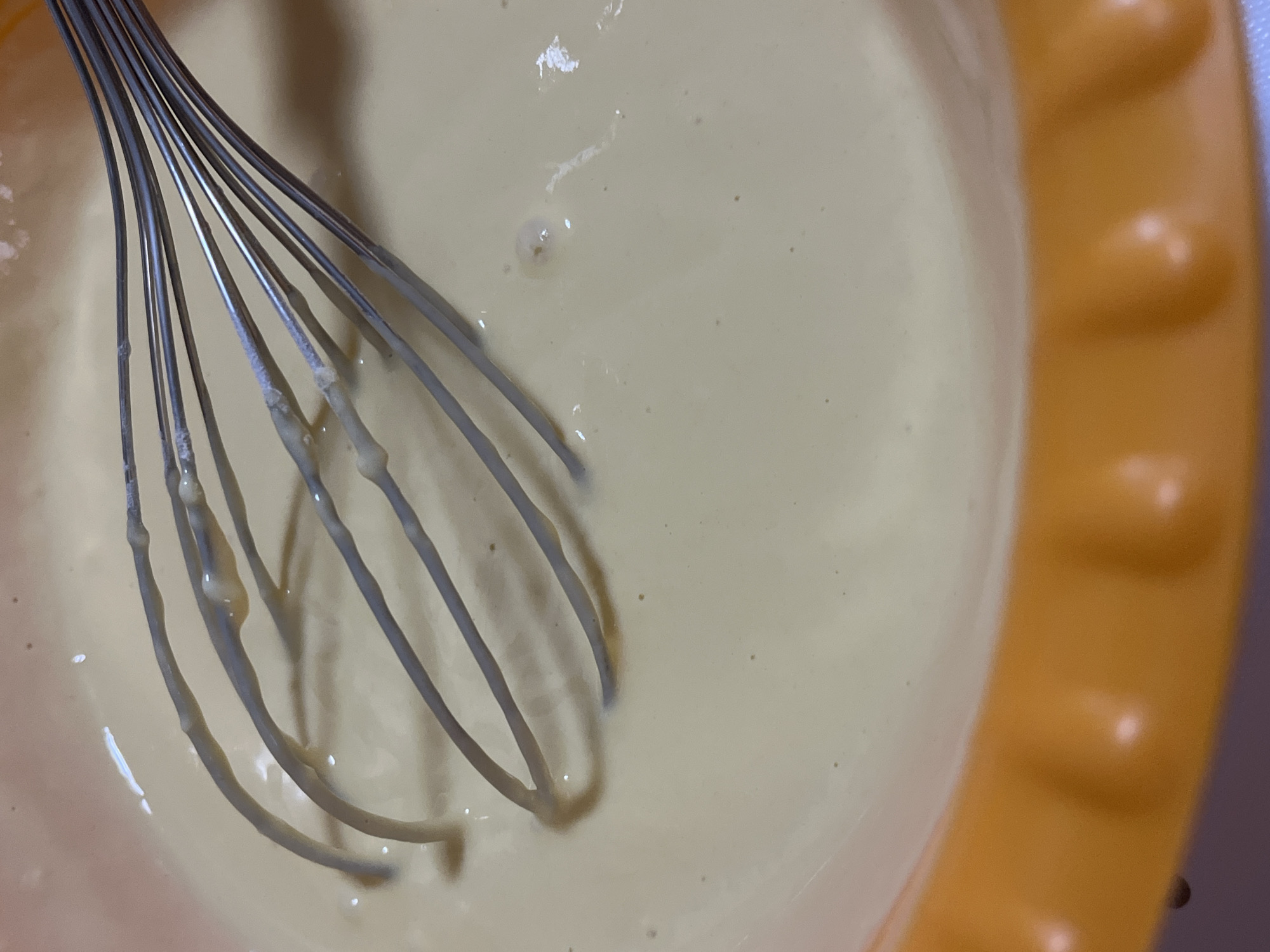 A close-up, overhead shot shows a whisk immersed in a light yellow batter inside an orange bowl. The batter has a smooth consistency with a few small bubbles visible. The whisk, made of silver metal, has multiple wires that are coated with some of the batter. The bowl has a textured rim with several indentations. The lighting suggests an indoor setting. The image does not contain information about the city of Krasnyye Barrikady, Russia.