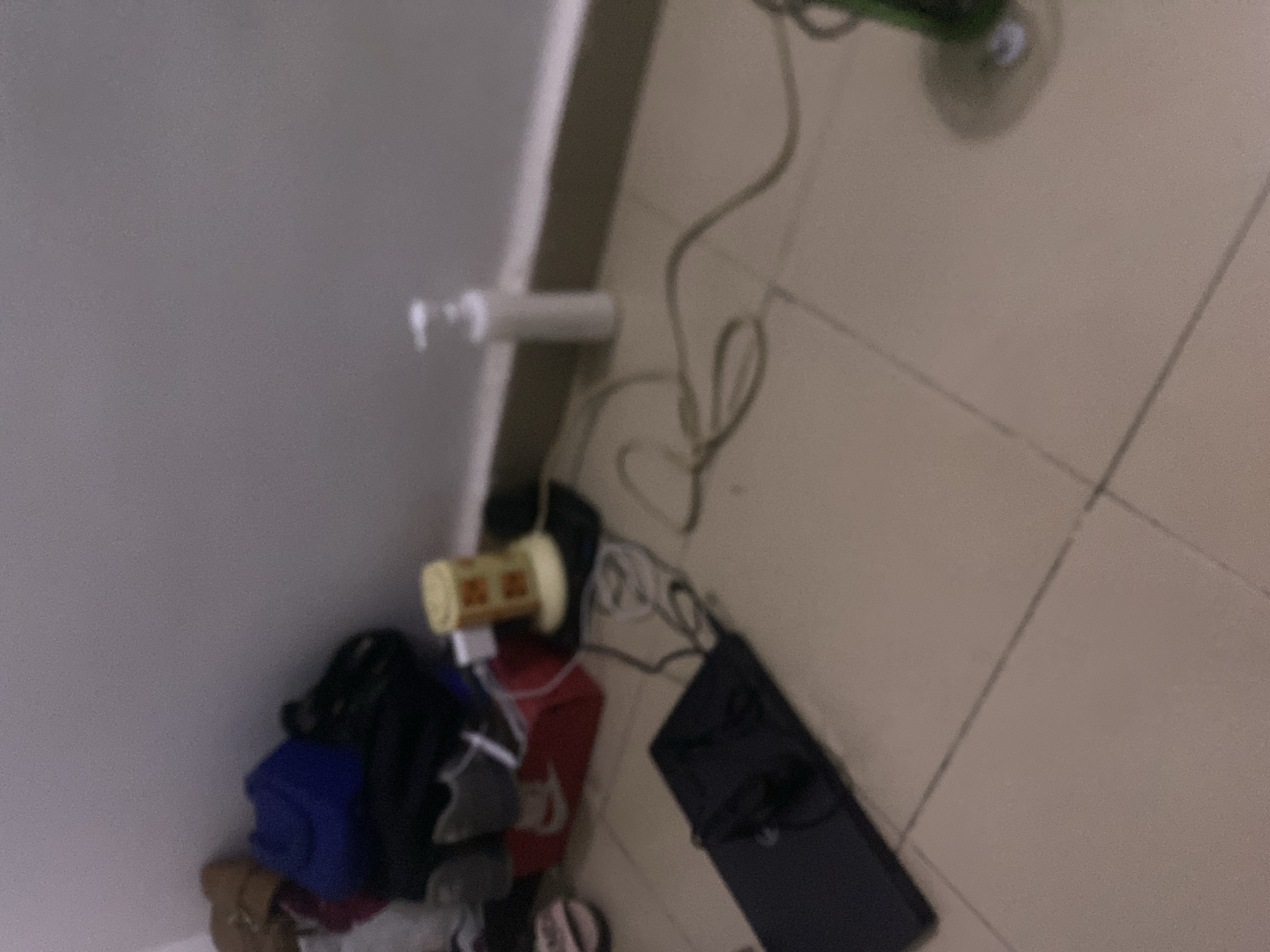 The image depicts a cluttered corner of a room, likely a bedroom or living space, on a tiled floor. In the upper left, a pile of items includes what appear to be shoes, a blue bag, and other dark-colored objects. Further right, a red box sits on the floor, with a yellow cylindrical object on top of it, connected to a black mat. To the right of this, a white pump bottle stands upright against the wall. In the foreground, a black flat object, possibly a laptop or tablet, is positioned on the floor, with dark cables trailing from it. Another coiled cable extends across the floor. The overall impression is one of disarray. The location is identified as Howayi, Nigeria.