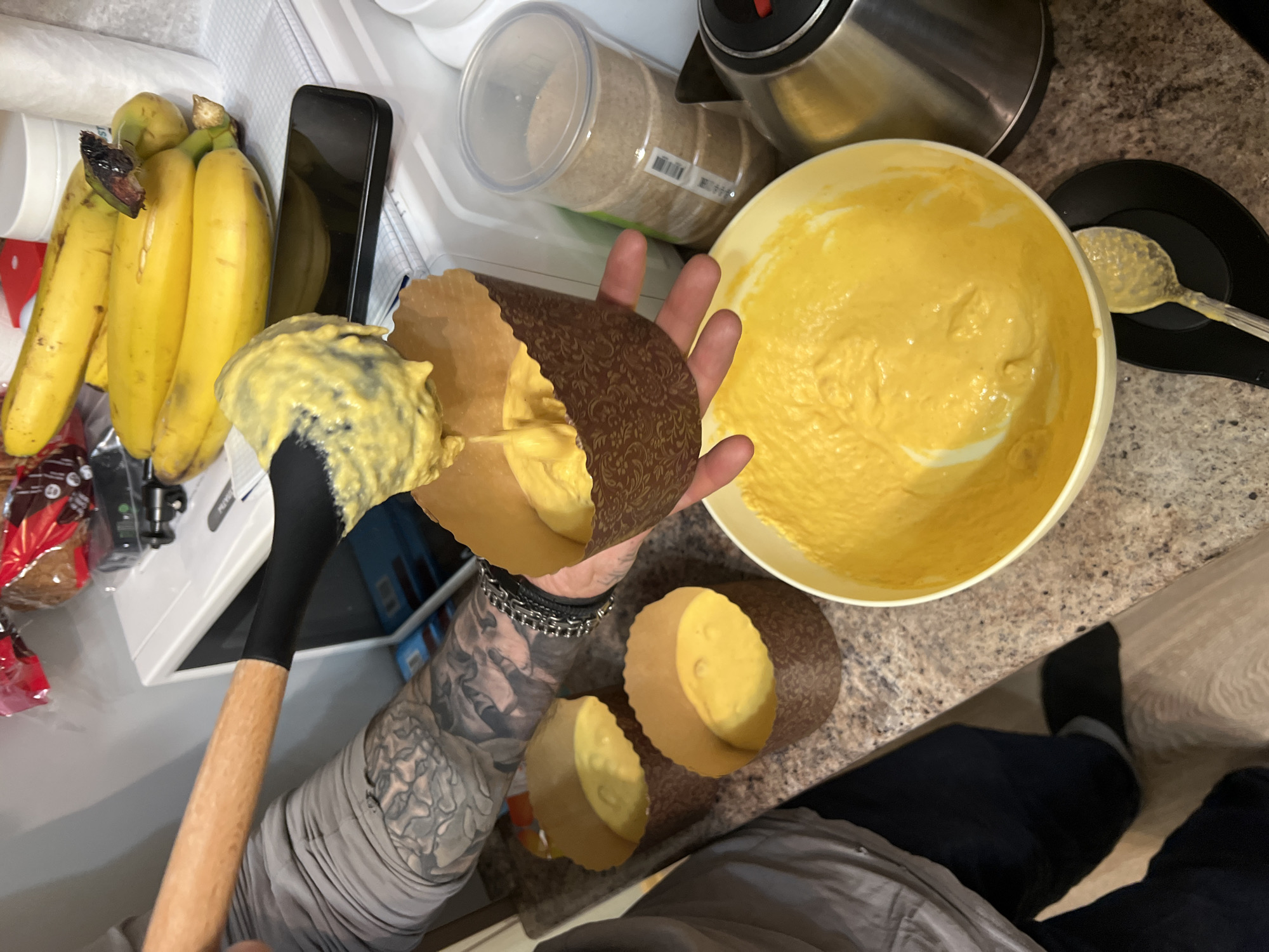 The image depicts a kitchen scene, focusing on the process of filling cupcake liners with a yellow batter. A person with tattooed arms is holding one liner and using a spatula to transfer batter from a large yellow bowl. Two other filled liners are visible on the countertop.
Nearby are a bunch of bananas, a smartphone, a clear container, and a stainless-steel kettle. The countertop has a speckled pattern. A spoon resting on a black spoon rest sits beside the bowl. The scene appears to be indoors. No information from the image indicates that this is in Saint Petersburg, Russia.