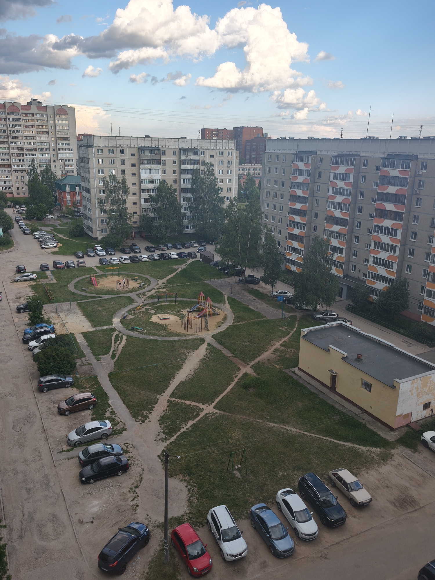 This is an aerial view of a residential courtyard in Yoshkar-Ola, Russia, during the daytime. The scene is dominated by large, multi-story apartment buildings typical of Soviet-era architecture. In the foreground, a paved area is lined with parked cars, including a dark blue hatchback, a red sedan, and a white SUV. Further into the courtyard, a patch of green grass is interrupted by dirt paths. Two playgrounds with swings and slides are visible, one with sand and climbing structures, and the other with a red slide. The courtyards are bordered by more apartment buildings, some with balconies and patterned facades. The sky above is blue with scattered white clouds. The overall impression is of a typical urban residential environment.
