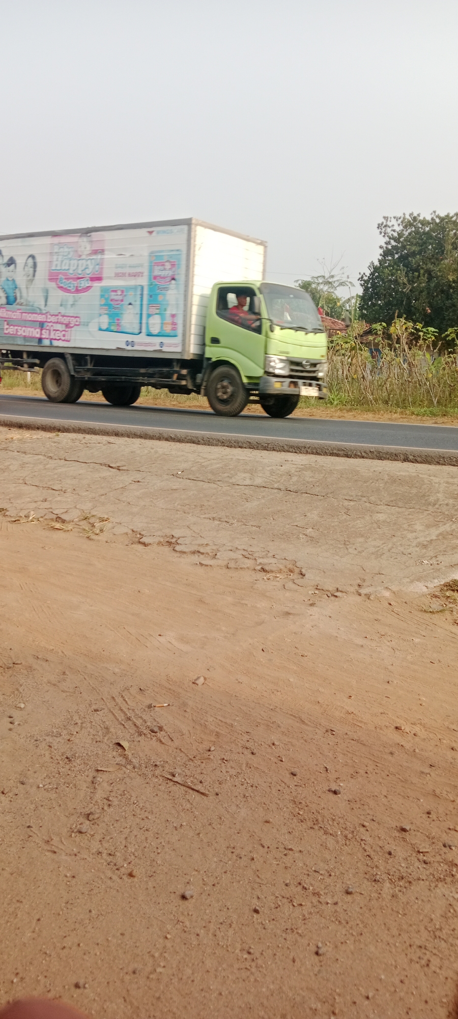 A light green truck with a white box body is driving on an asphalt road. The truck is decorated with a banner that says "Happy! Baby" and features images of babies and diapers. The truck is in motion, as indicated by the blurred wheels. The driver of the truck is visible through the windshield, wearing a red shirt. The background consists of trees, bushes, and a clear sky. The foreground shows a dirt road with some cracked asphalt sections. The scene appears to be in a rural or semi-rural area, possibly a highway or main road. The location is specified as Gondang Rejo, Indonesia.