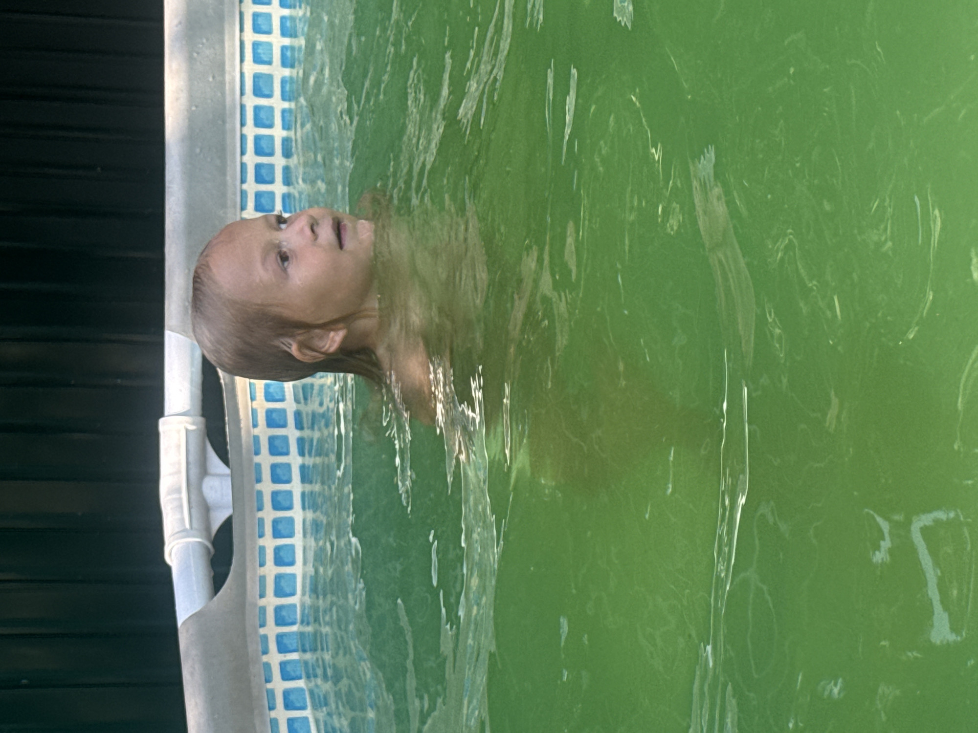In this media file, a young child is partially submerged in green water, likely a swimming pool due to the presence of a blue tiled liner and a white PVC pipe structure along the edge. The child's head and shoulders are visible above the water, with their face turned upwards and to the left, appearing to be looking at something or someone. Their expression is somewhat neutral, with an open mouth. The water is murky and green, suggesting it may not be clean or properly treated. The background shows the dark, horizontal slats of what appears to be a fence or wall. The image is oriented vertically. The setting suggests a sunny day, typical for outdoor pool activities, though no specific weather details are discernible. The location context provided is Sovietskyi, Ukraine. No text is visible in the image.